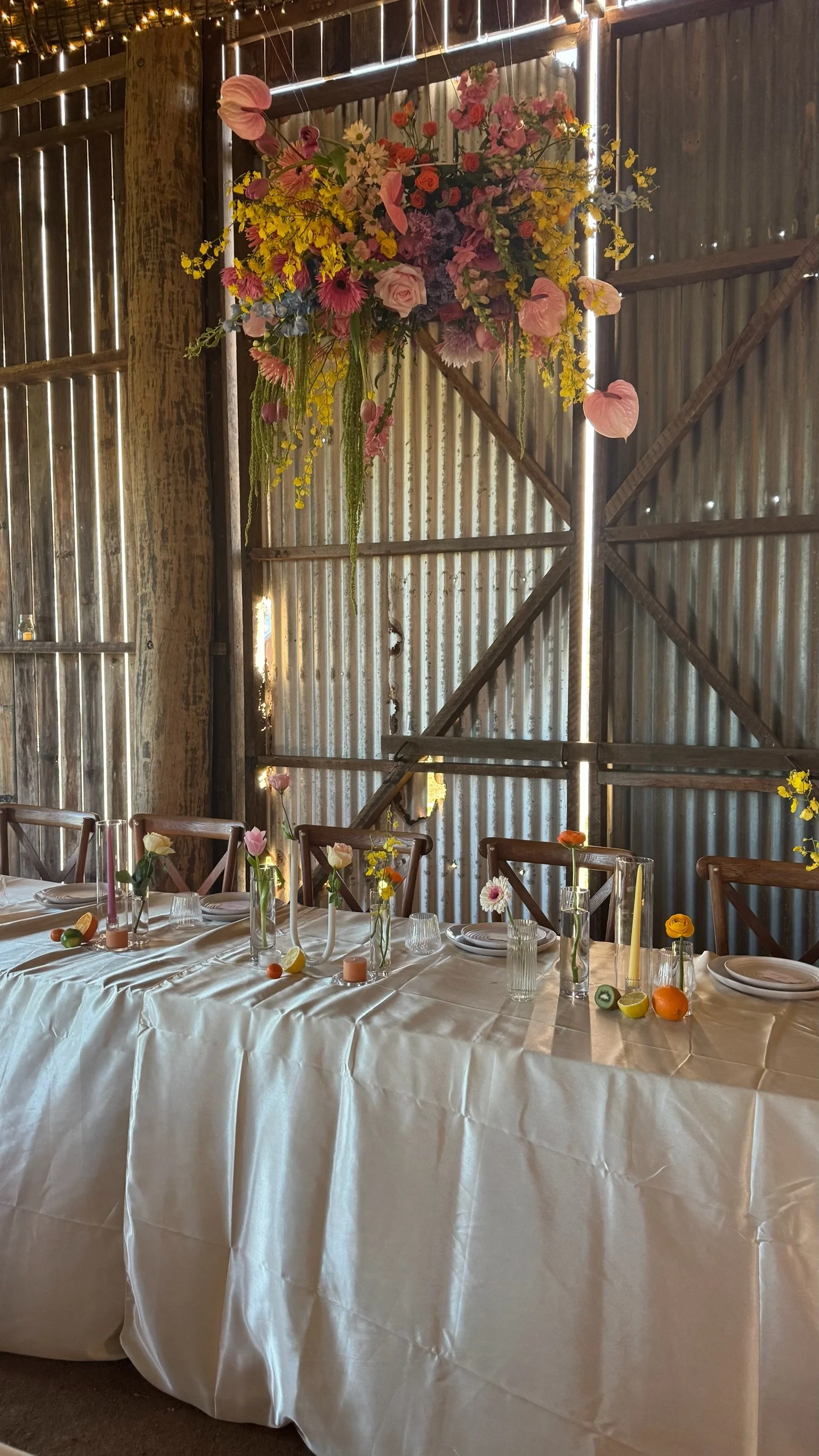 A rustic wooden barn with a long table set with a white tablecloth, small glass vases with colorful flowers, and fruit slices as decorations. A large floral arrangement hangs on the wall behind the table.