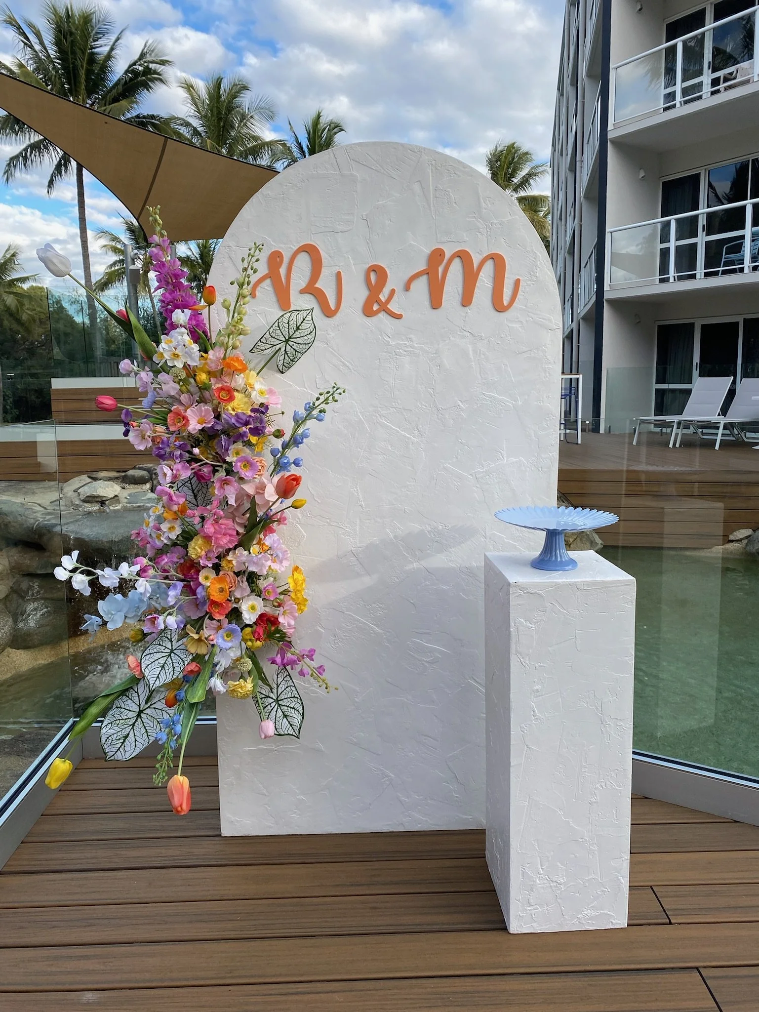 Decorative display with a white textured backdrop featuring the initials R & M in orange cursive letters, a colorful floral arrangement on a white pedestal, and a blue pedestal with a decorative dish, set on a wooden deck near a pool with a resort bu