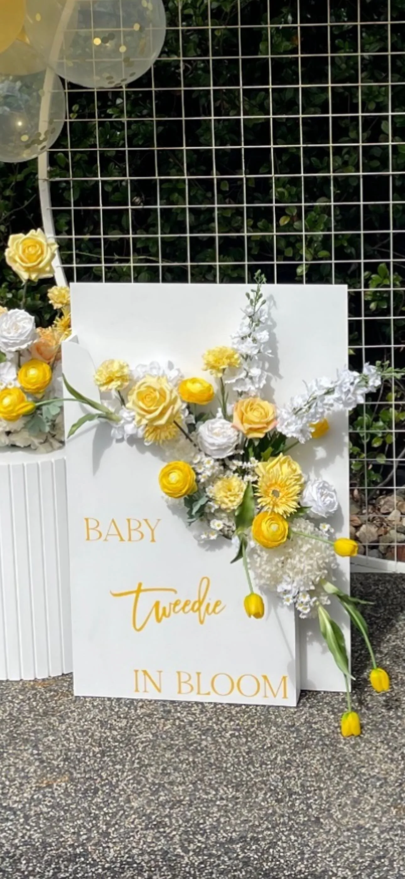 Floral arrangement with white and yellow flowers next to a sign that reads 'Baby Tweedie in Bloom' at a celebration event.