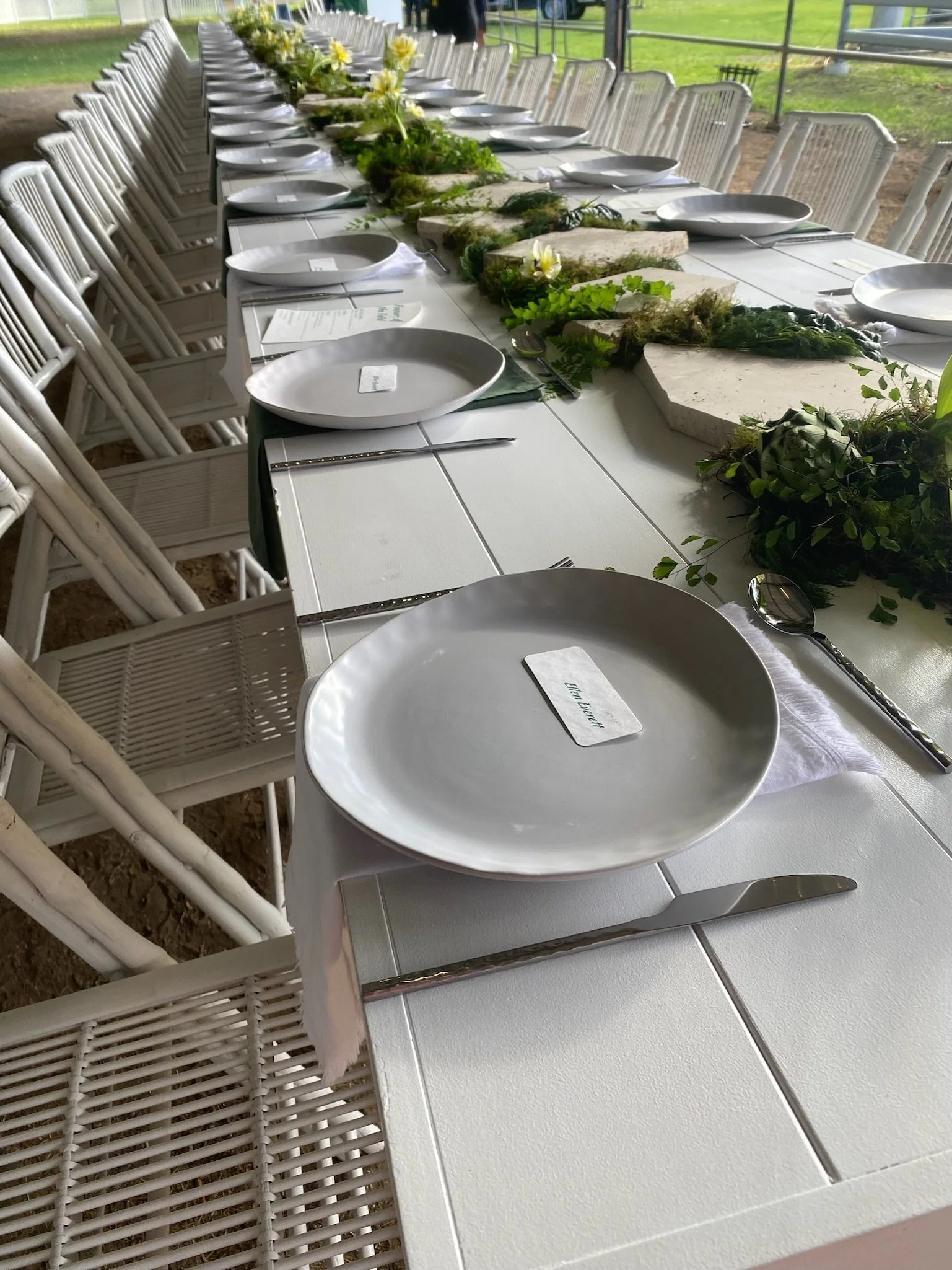 A long dining table set outdoors with white plates, silverware, and a green centerpieces with flowers and greenery.