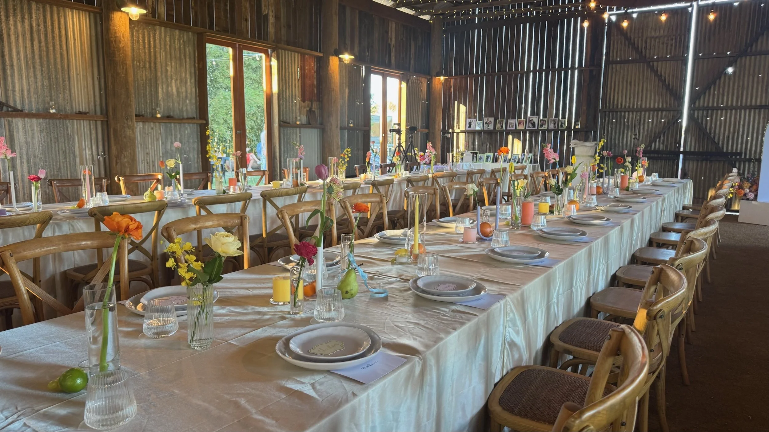 Long dining table set for a celebration with colorful flowers, candles, and plates in a rustic barn with wood and metal walls.