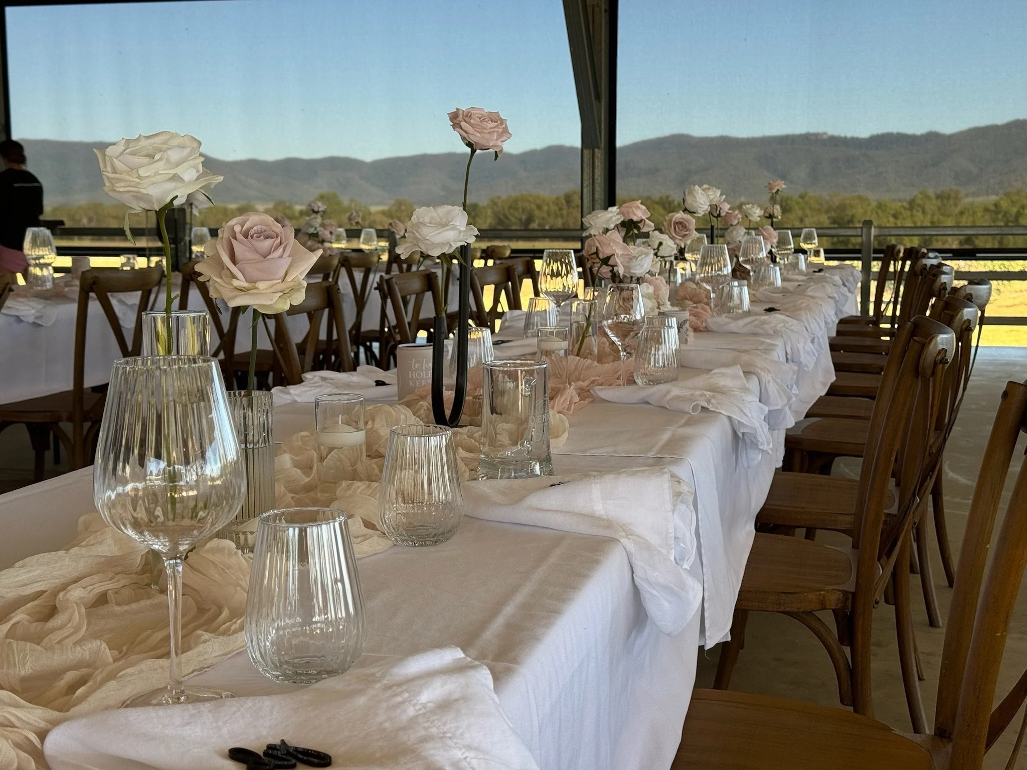 Elegant outdoor dining table decorated with blush and white roses, glassware, and candles, set against a scenic mountain view.