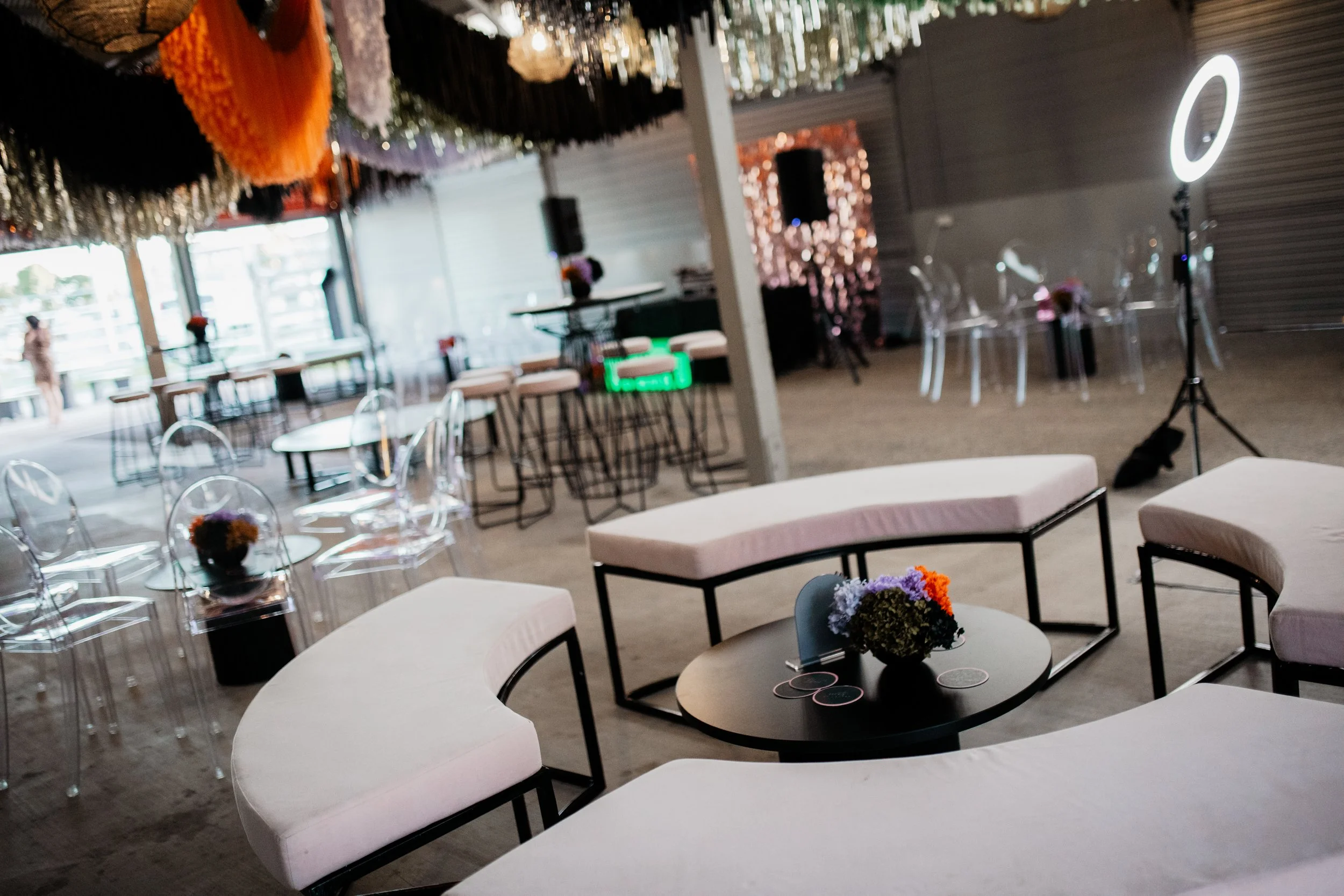 Interior of a decorated event space with modern furniture, floral centerpieces, a ring light, and colorful hanging decorations.