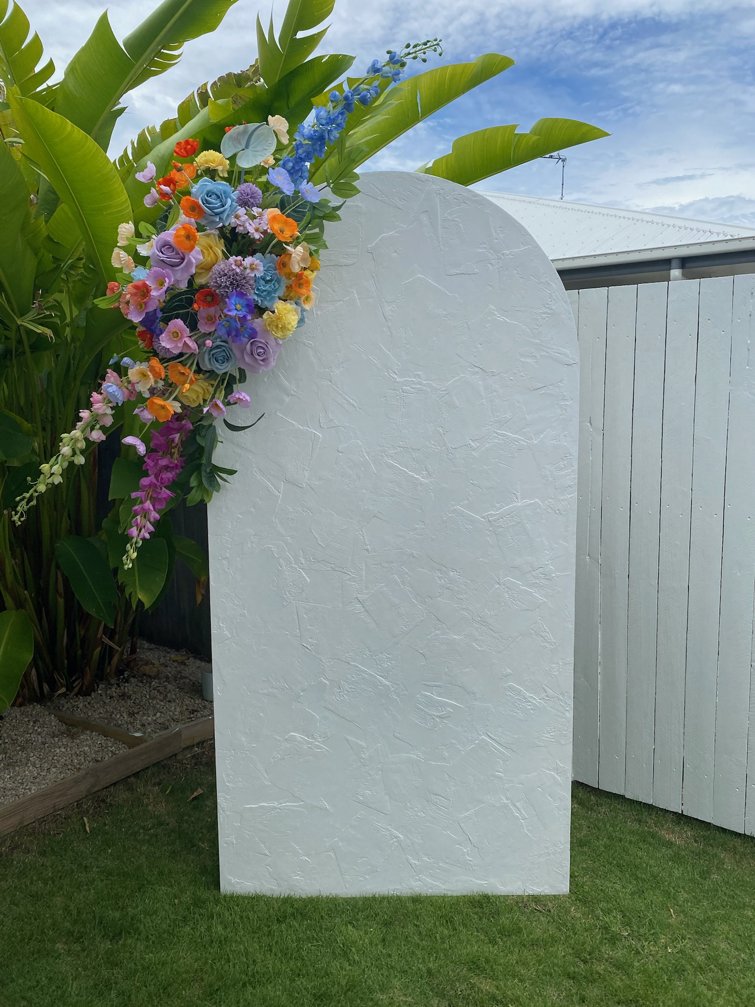 A white textured stone memorial with a floral arrangement on the upper left corner, set outdoors with green grass, tropical plants, a white picket fence, and a cloudy sky in the background.