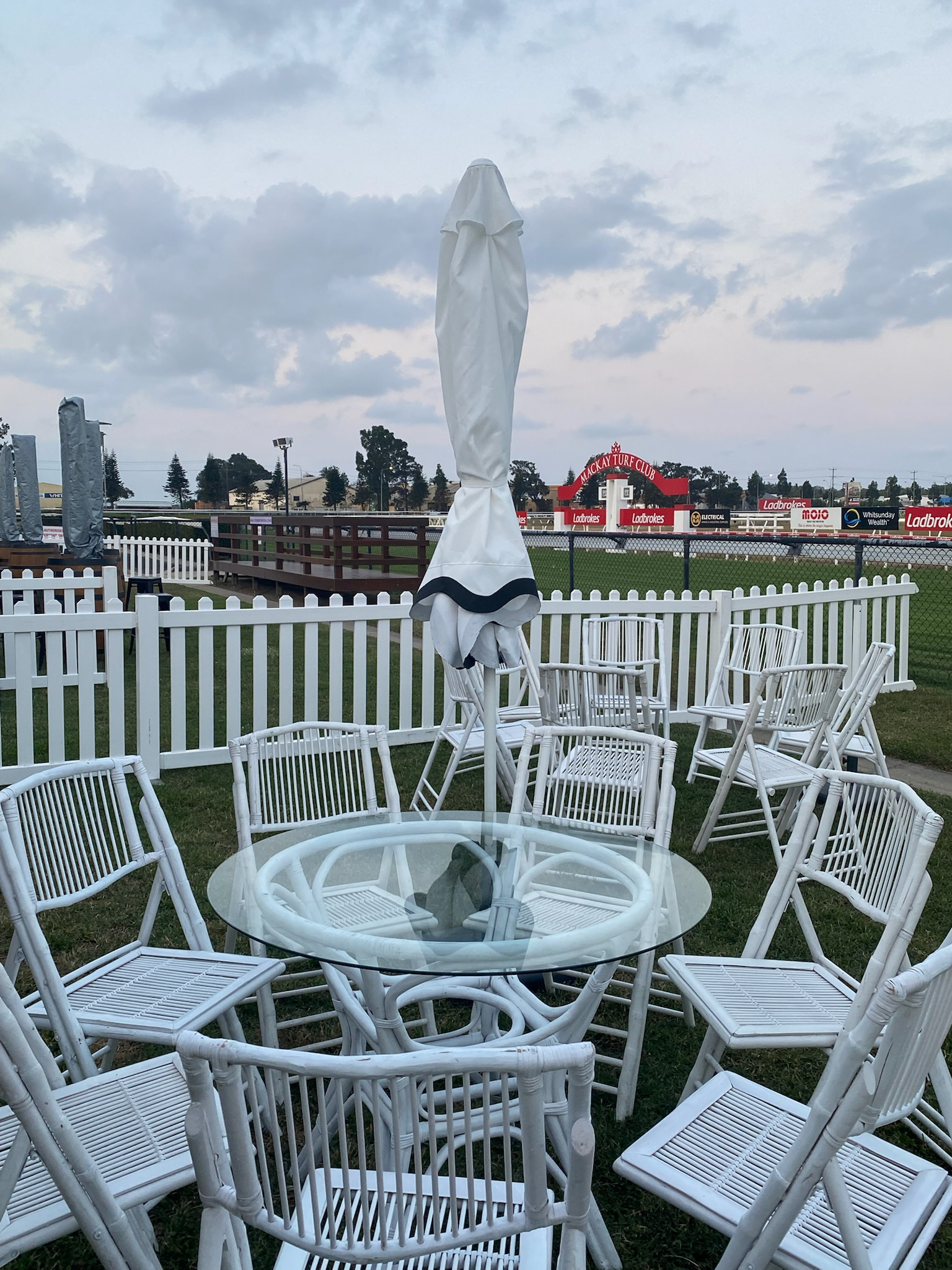 Empty outdoor seating area with white chairs and a round glass table, with an umbrella in the center, on a grassy area enclosed by a white picket fence, nearby a sports field with advertisements, under a cloudy sky.