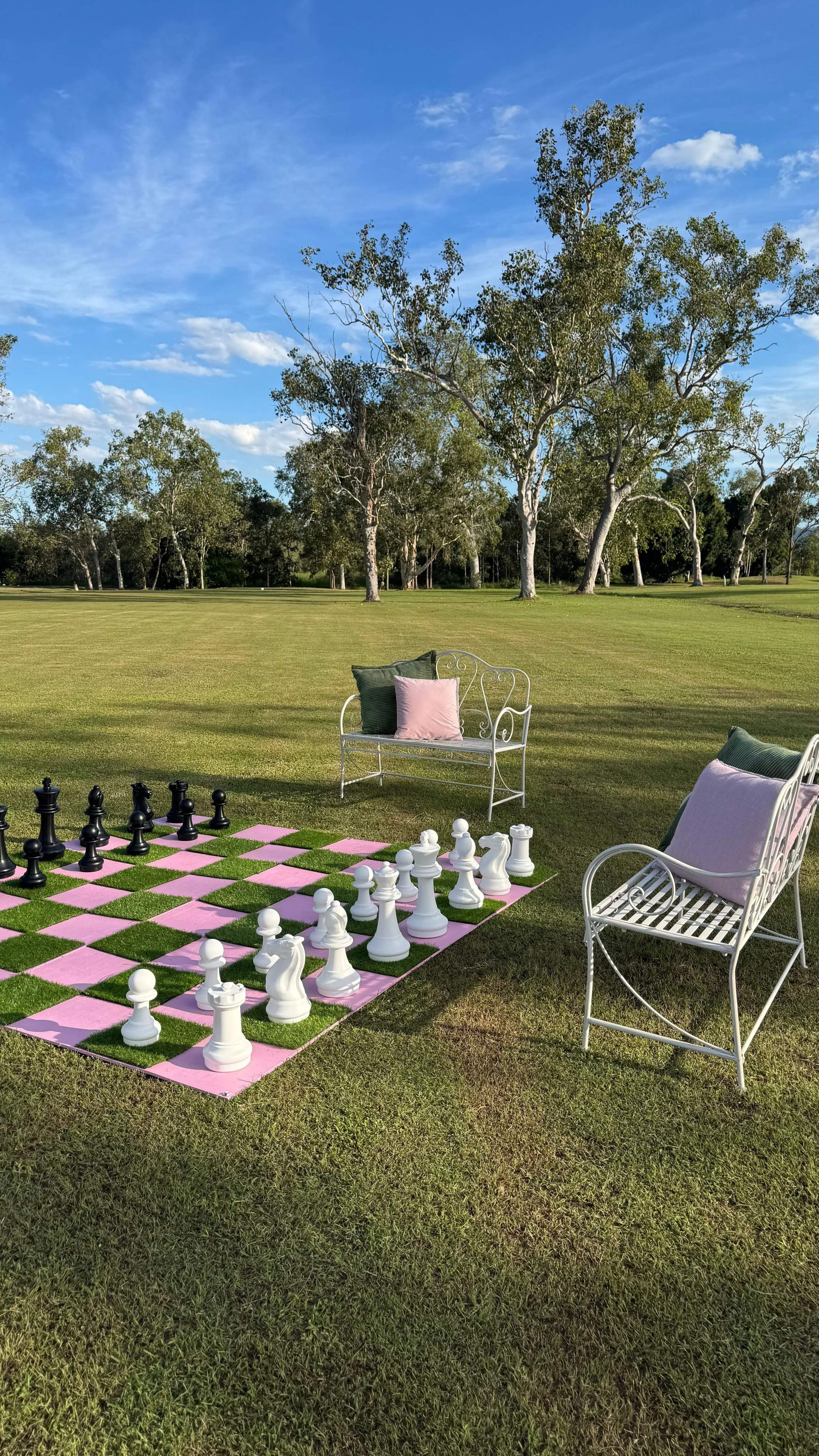 A large outdoor chessboard on grass with white and black chess pieces. Two white metal benches with pastel cushions and pillows are nearby. Trees and a blue sky with clouds in the background.