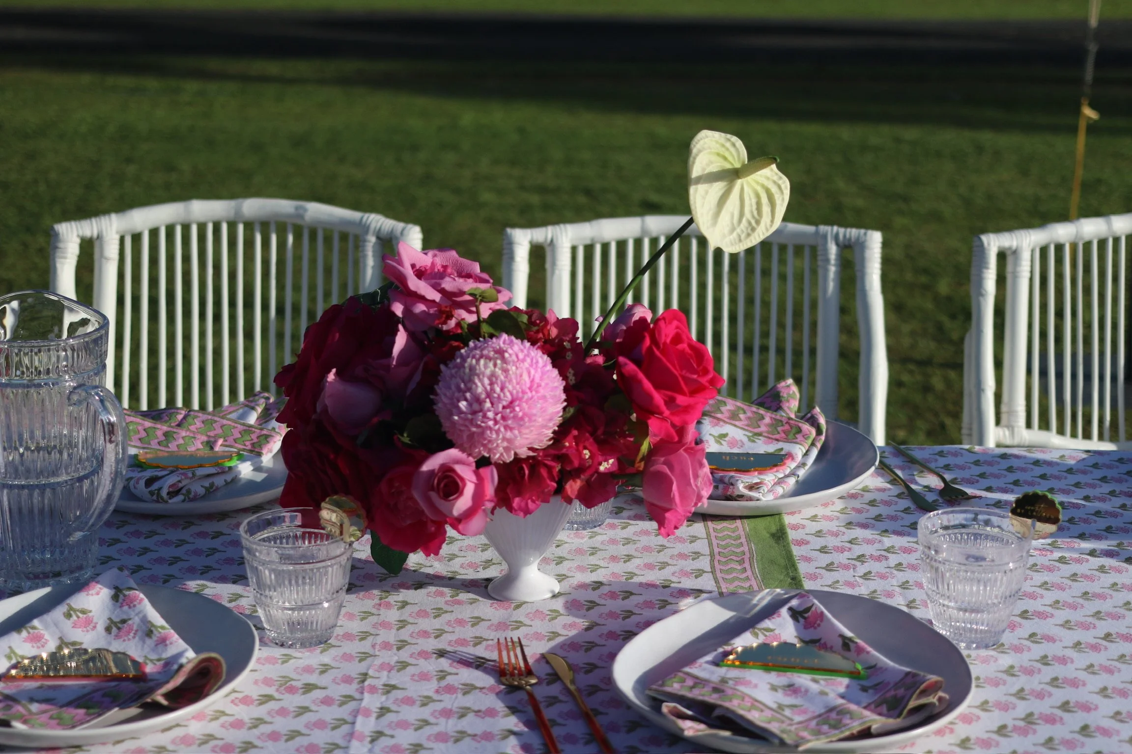 A table set for an outdoor event with a floral centerpiece featuring pink and white flowers, including roses and chrysanthemum, on a white and pink floral tablecloth. There are white plates with colorful napkins, clear glassware, and silverware, and 
