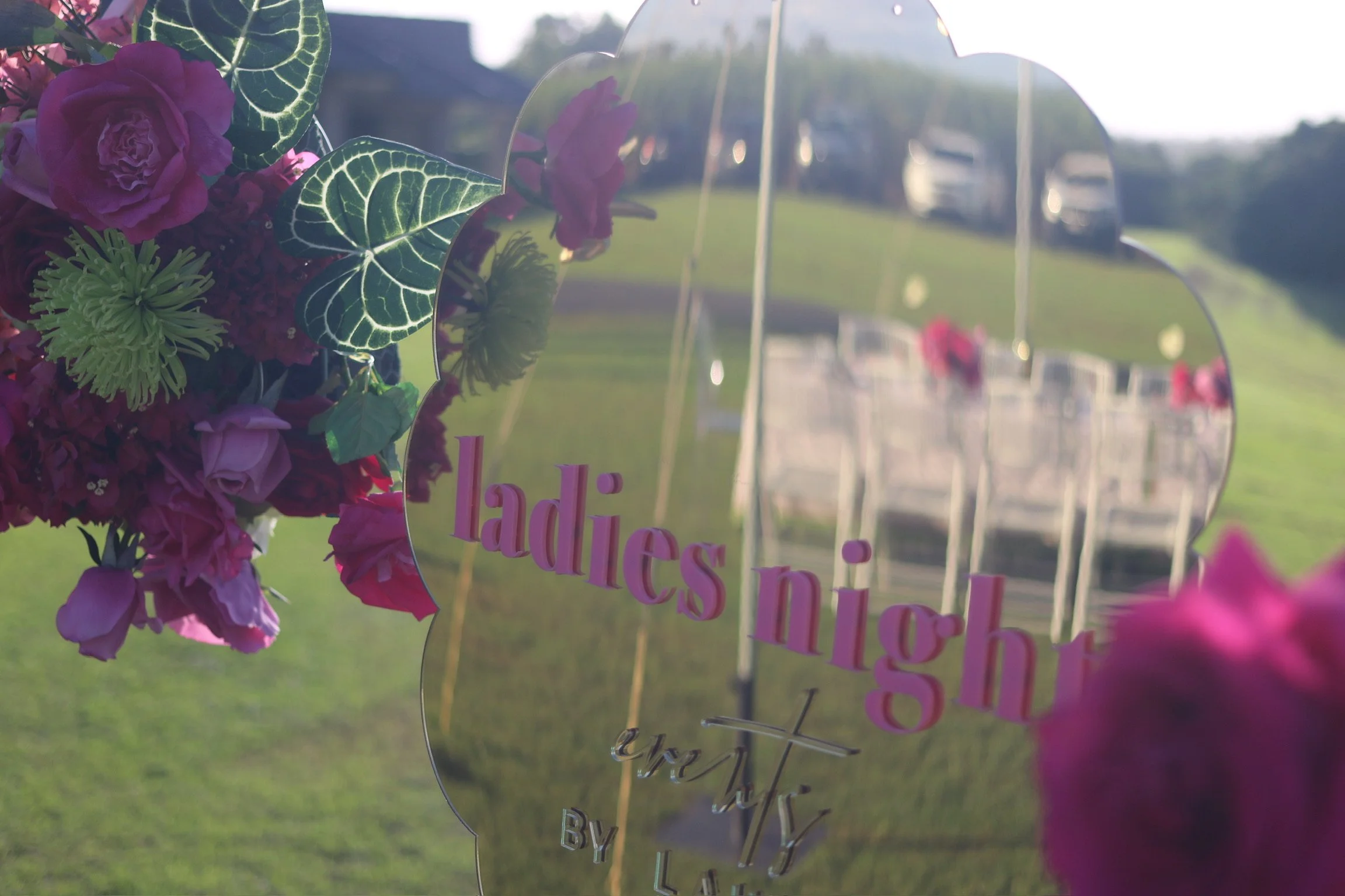 Metal heart-shaped wedding sign with pink and purple flowers on the left, with the reflection of chairs and car in the mirror surface of the sign, standing on a grassy outdoor area.