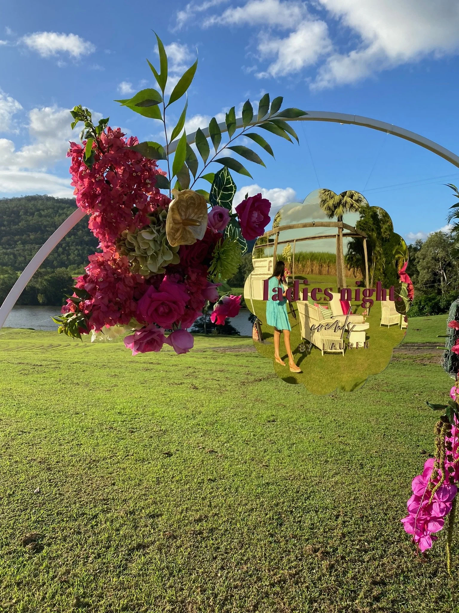 Decorative mirror and floral arrangement outdoors on a grassy field with hills and sky in the background, with a sign reading 'Ladies Night'