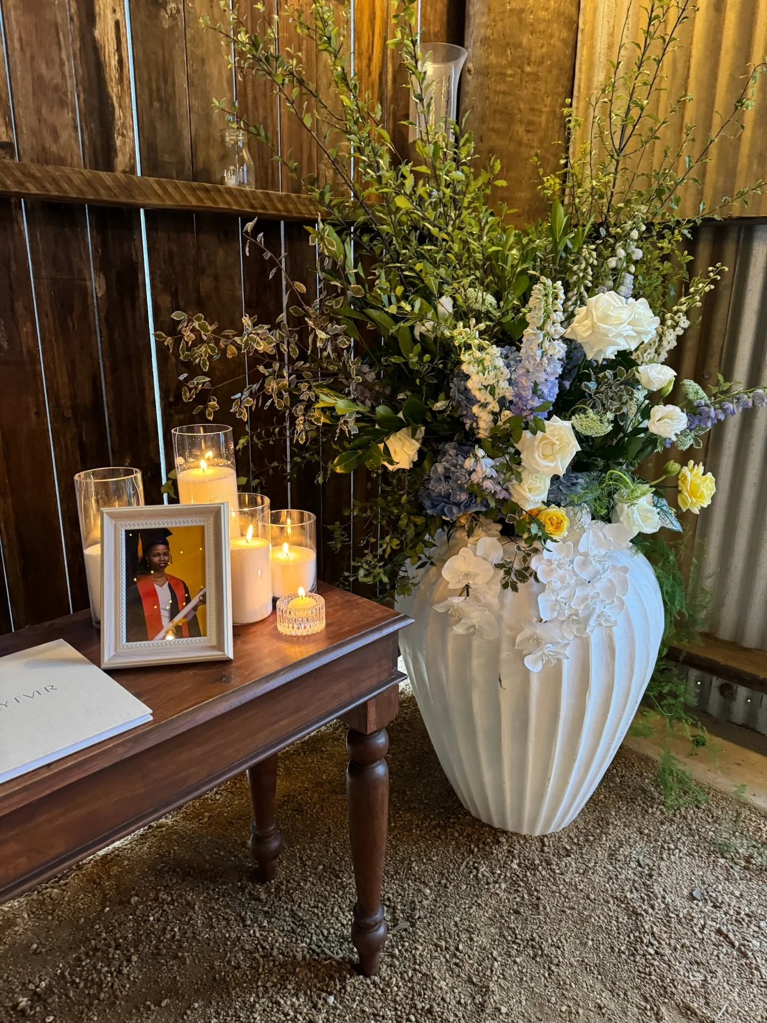 Large floral arrangement in a white vase, with white, purple, and green flowers and foliage. Small lit candles and a framed photo of a woman in graduation attire are on a wooden table beside the arrangement, against a wooden wall.