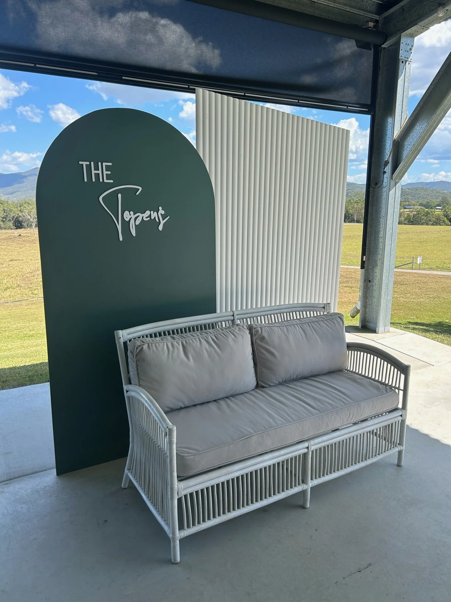 Outdoor seating area with a white rattan sofa with gray cushions, positioned against a green backdrop with white text that says 'The Taper' at an open venue overlooking a grassy field and distant mountains.