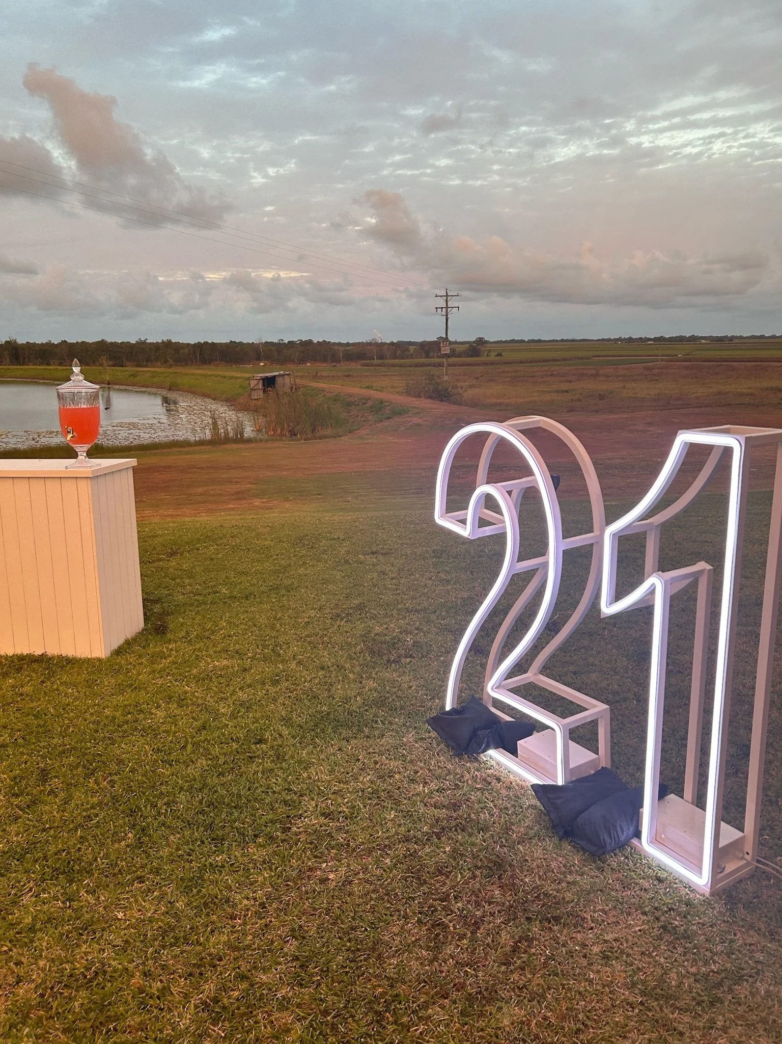 A large illuminated number 21 sign outdoors on grass, with a small table holding a drink in the background, a pond, and open field scenery under a cloudy sky.