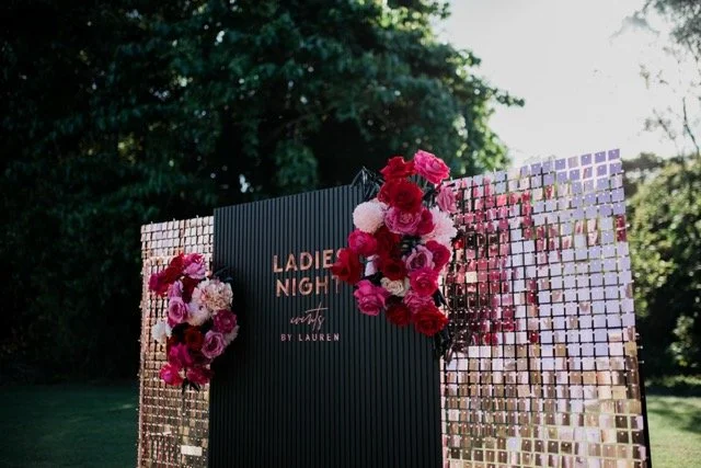 A decorative outdoor event backdrop with pink and white floral arrangements, gold mirror panels, and a black sign that reads "Ladies Night" by Lauren, set against a backdrop of green trees and sunlight.