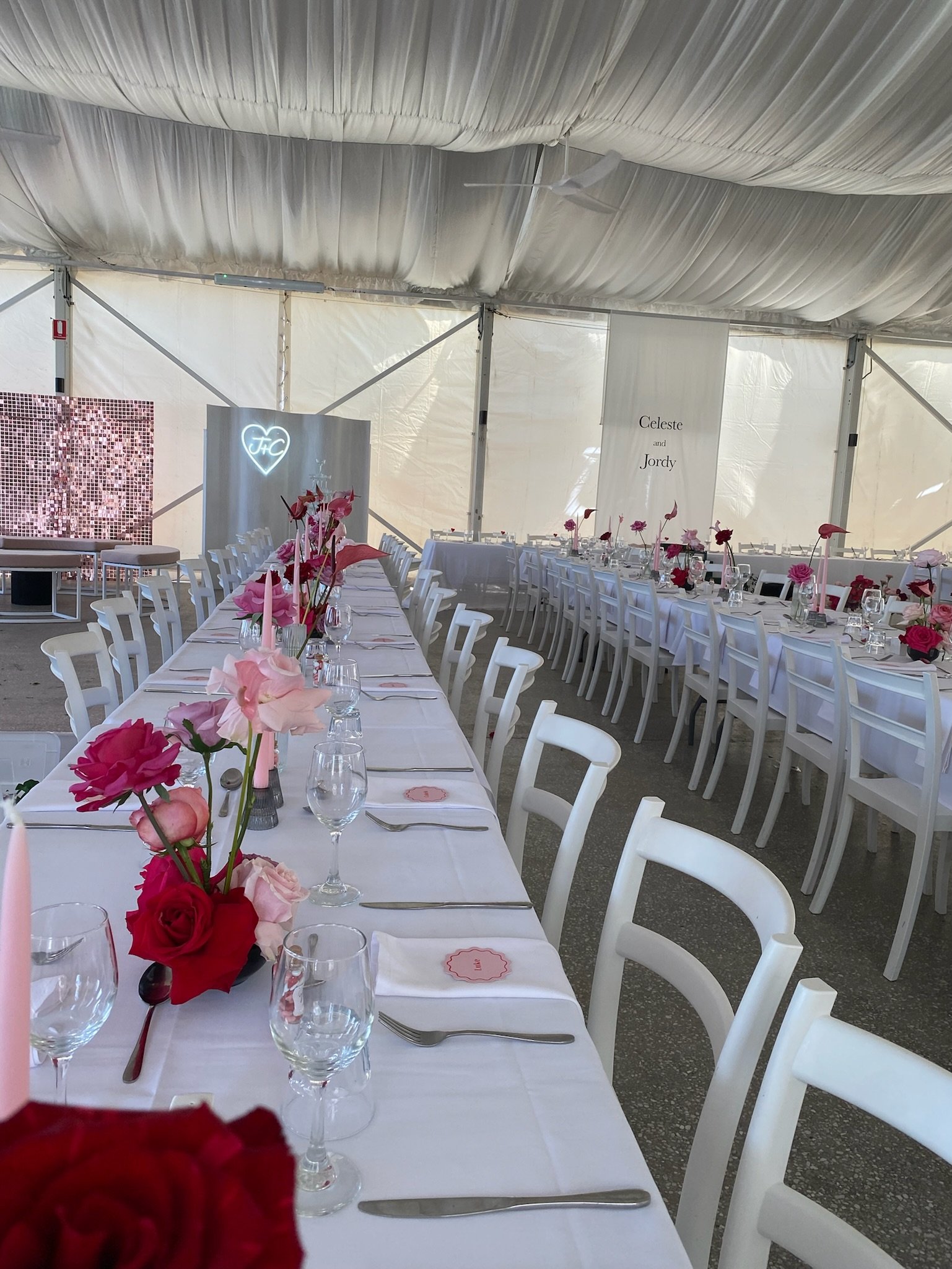 Wedding reception setup inside a white tent with long tables decorated with pink and red flowers, glassware, and place settings.