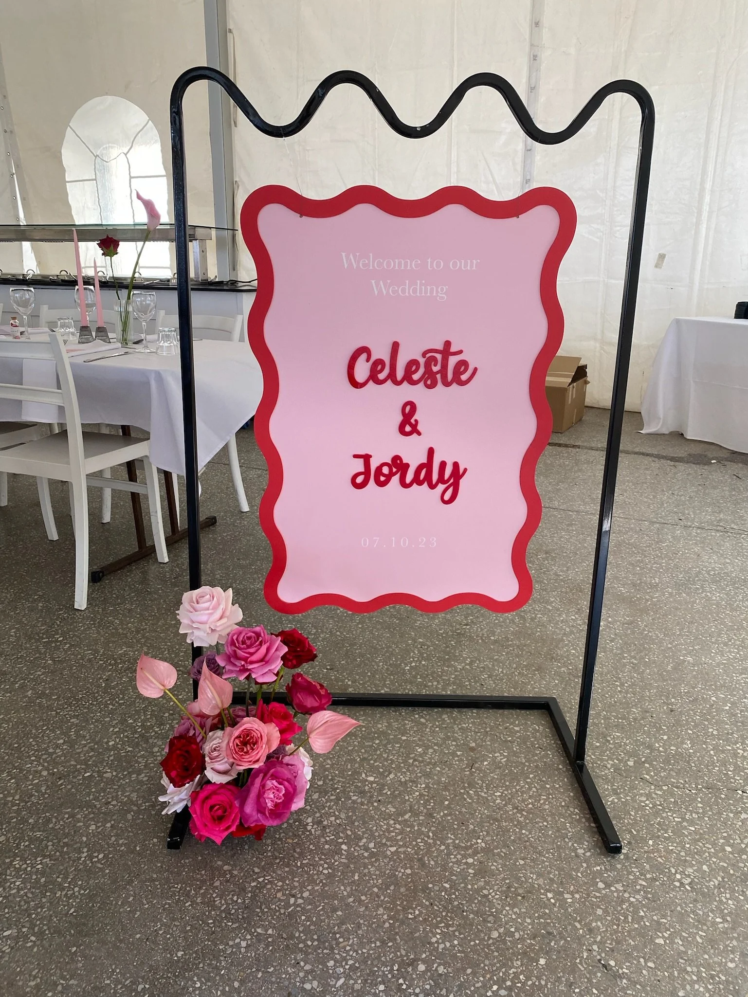 Wedding welcome sign with pink background and red border, displaying the names Celeste and Jordy, and the date 07.10.23, adorned with a bouquet of pink and red roses at the base in a tented reception area.
