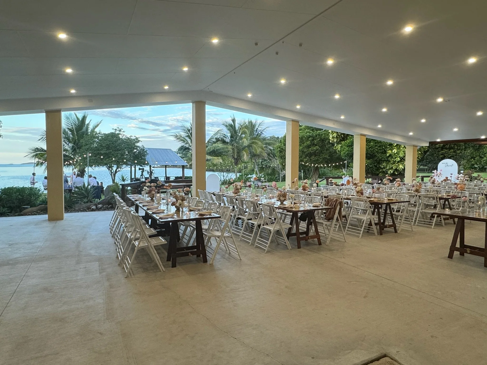 A spacious outdoor event area with tables and chairs arranged for a gathering or celebration. The setting includes a view of palm trees and the ocean in the background, with a partly cloudy sky and some guests near the water. The indoor ceiling is de
