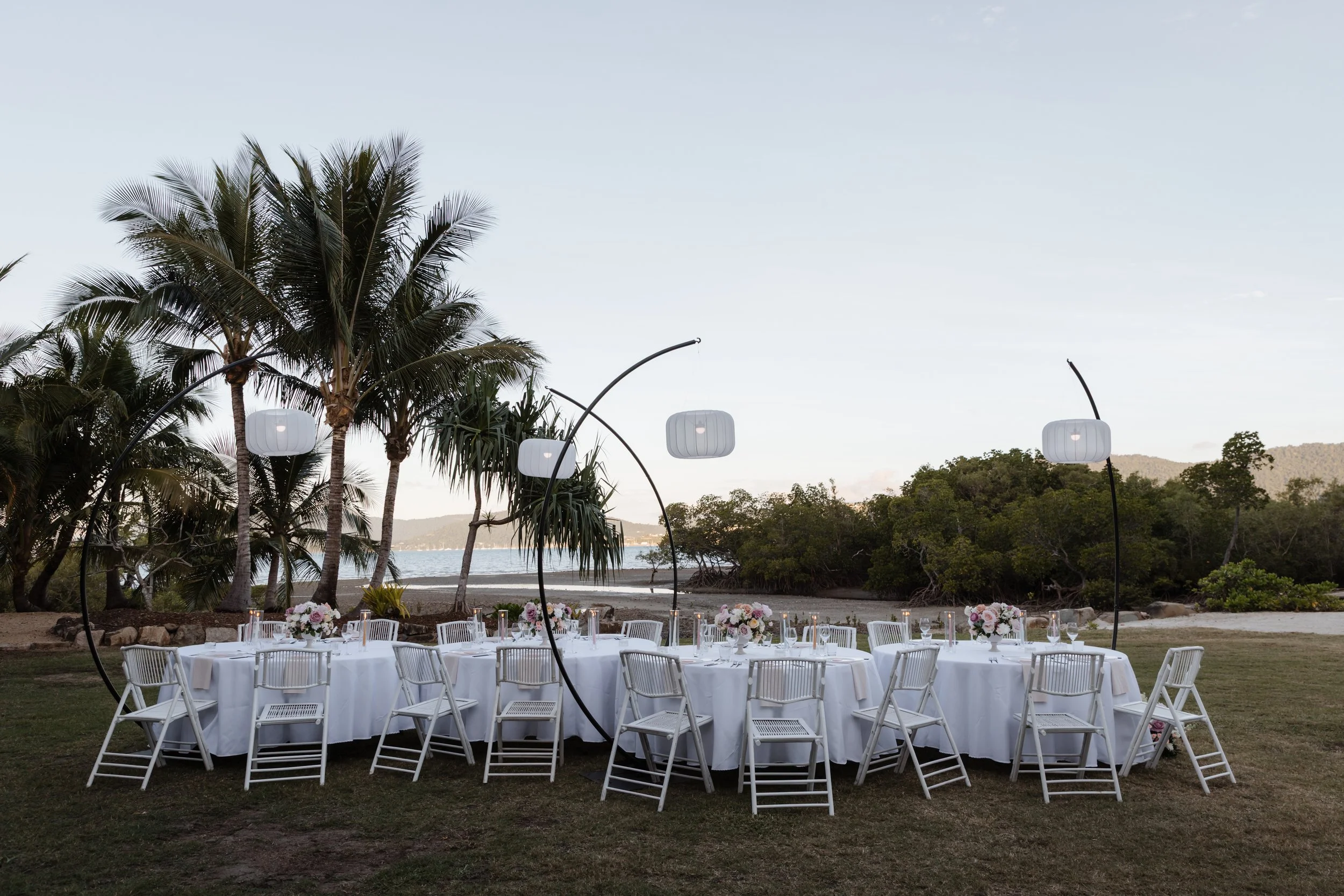 Outdoor seaside event setup with round tables, white tablecloths, floral centerpieces, white chairs, and modern hanging lanterns under tall palm trees and on a grassy area near the water.