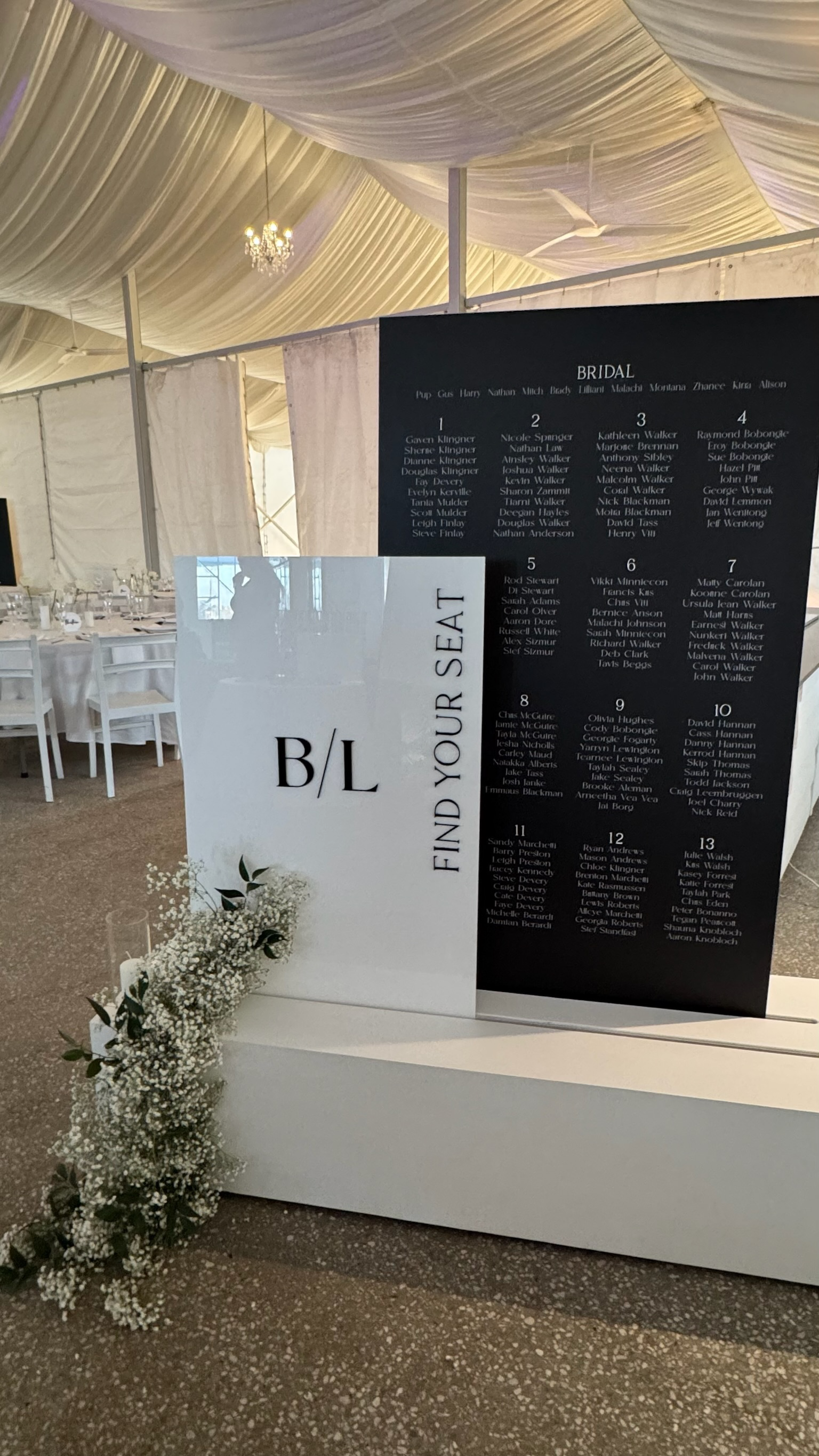 Wedding reception seating chart in a decorated event tent, with a sign that says 'FIND YOUR SEAT' and numbered tables listed.