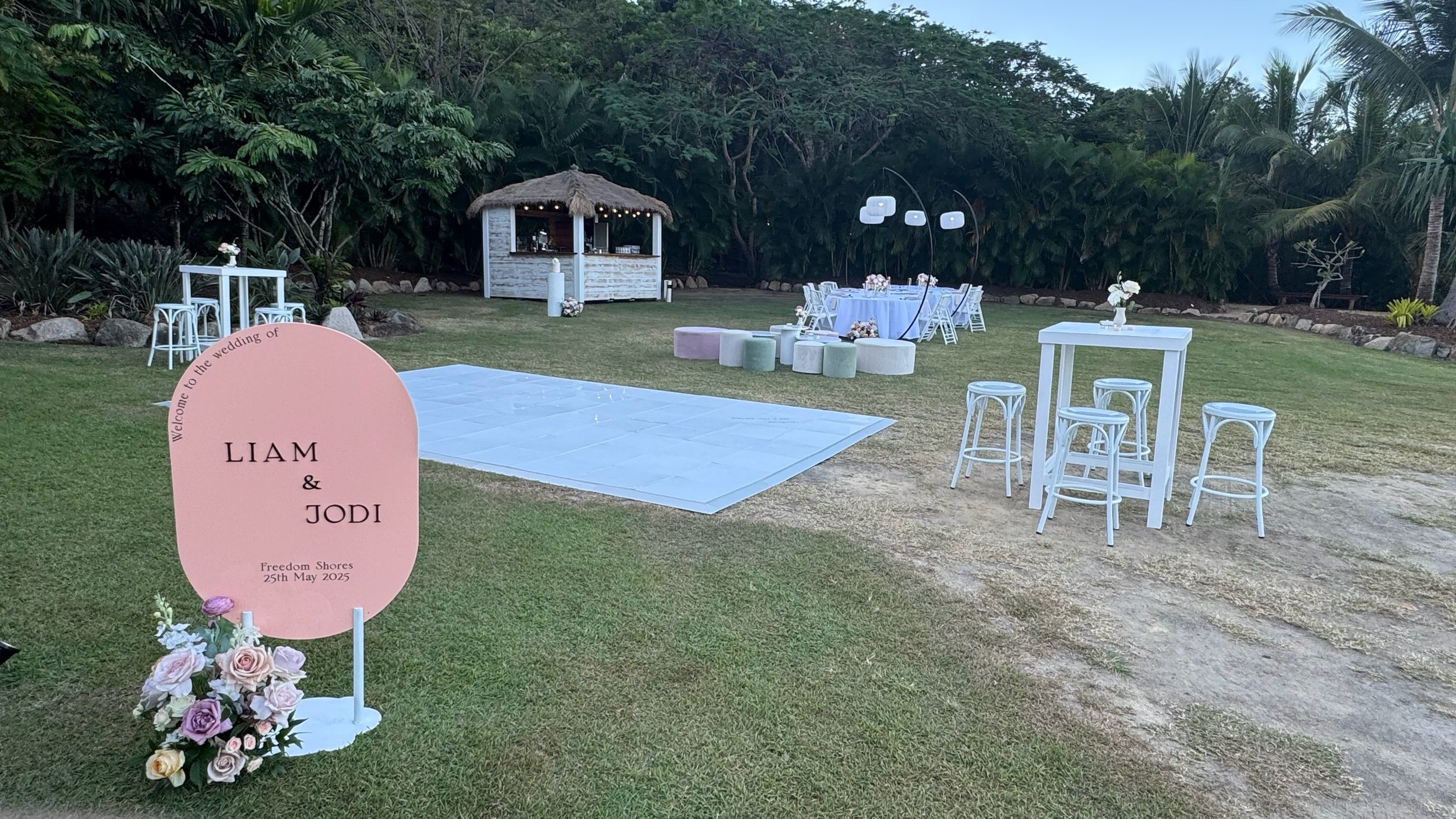 Outdoor wedding setup with a pink sign reading "Liam & Jodi, Freedom Shores, 25th May 2025" surrounded by flowers. There are white tables and stools, a dance floor, and decorative lighting in a garden with lush greenery and trees.