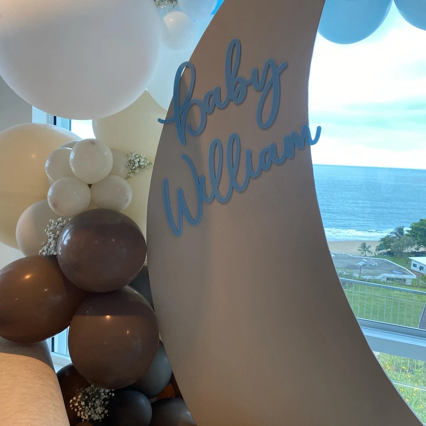 Decorative display with a large greeting sign reading 'baby William' in blue cursive, surrounded by white, cream, brown, and beige balloons, with a view of the ocean and beach through a window in the background.