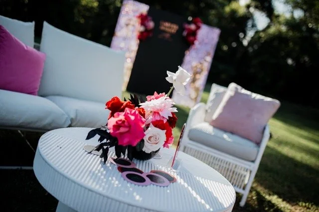 Outdoor setting with white wicker chairs with pastel cushions, and a white round table with a bouquet of pink, red, and white flowers and a pair of pink sunglasses.