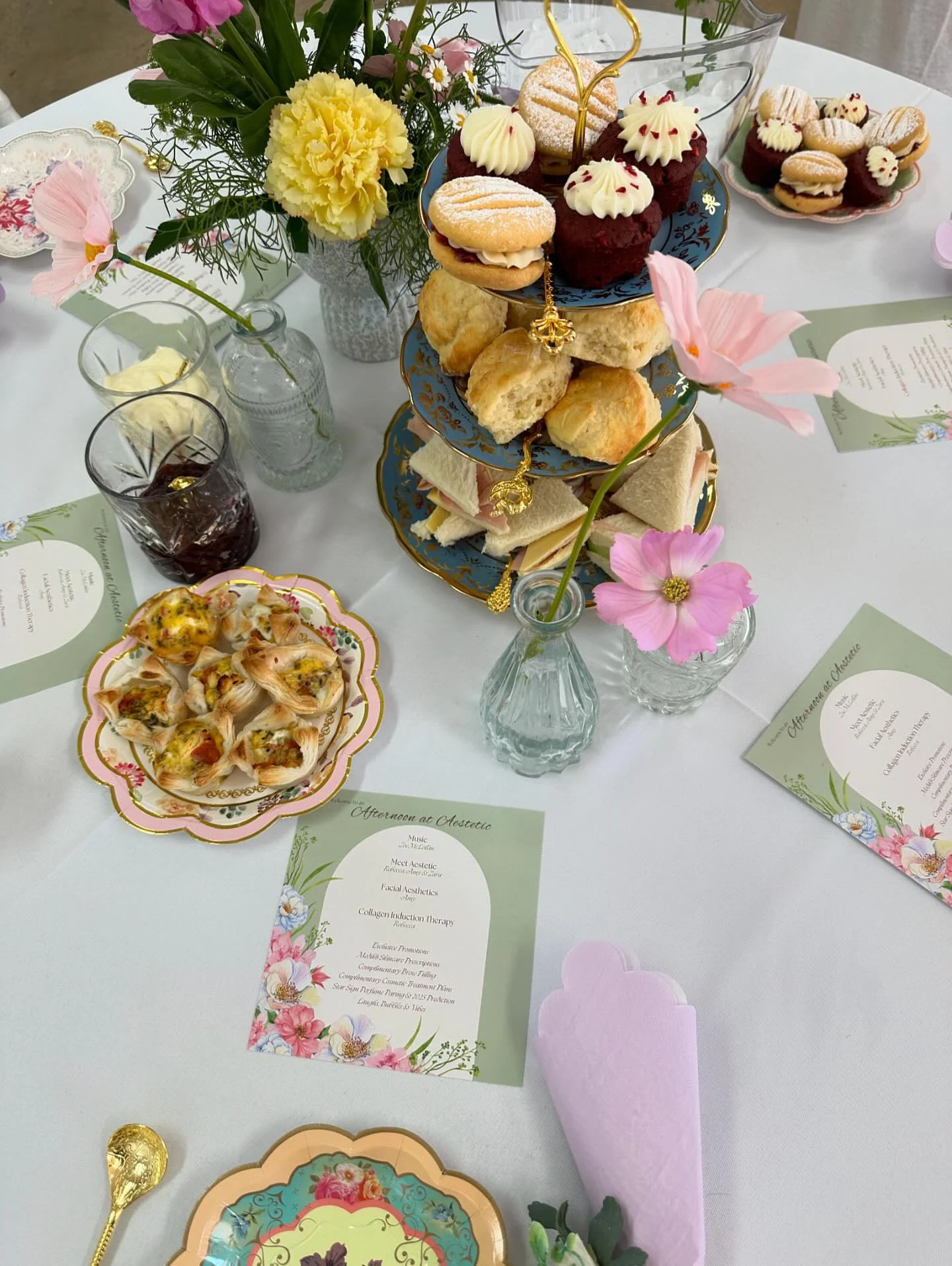 A table set for an afternoon gathering with a three-tier serving tray filled with assorted desserts, including scones, cupcakes, and sandwiches. The table is decorated with pink and yellow flowers in small vases, and there are individual plates of ap