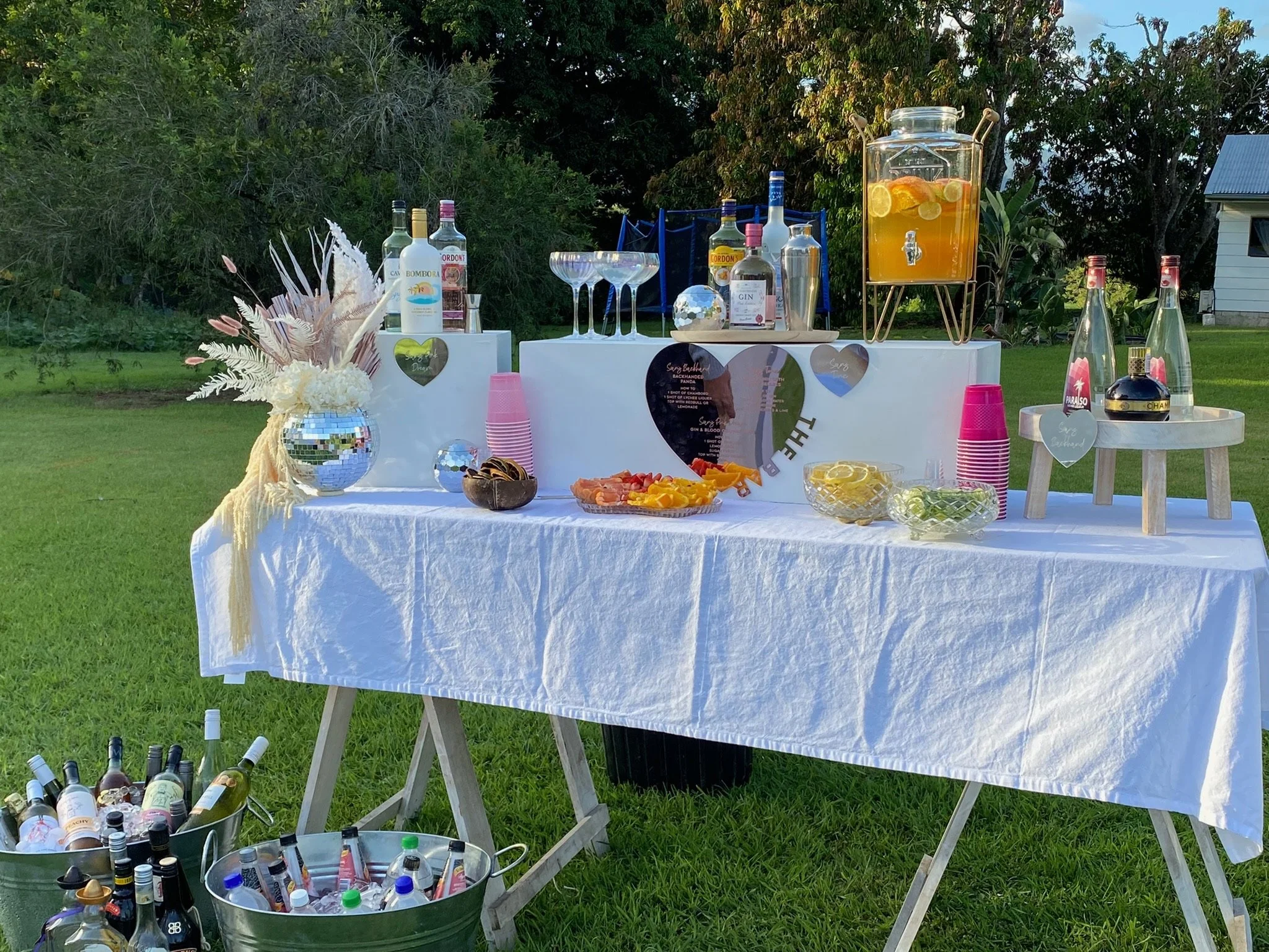 Outdoor party refreshment table with drinks, snacks, and decorations on a white tablecloth, surrounded by buckets of bottles on a grassy area with trees and a house in the background.