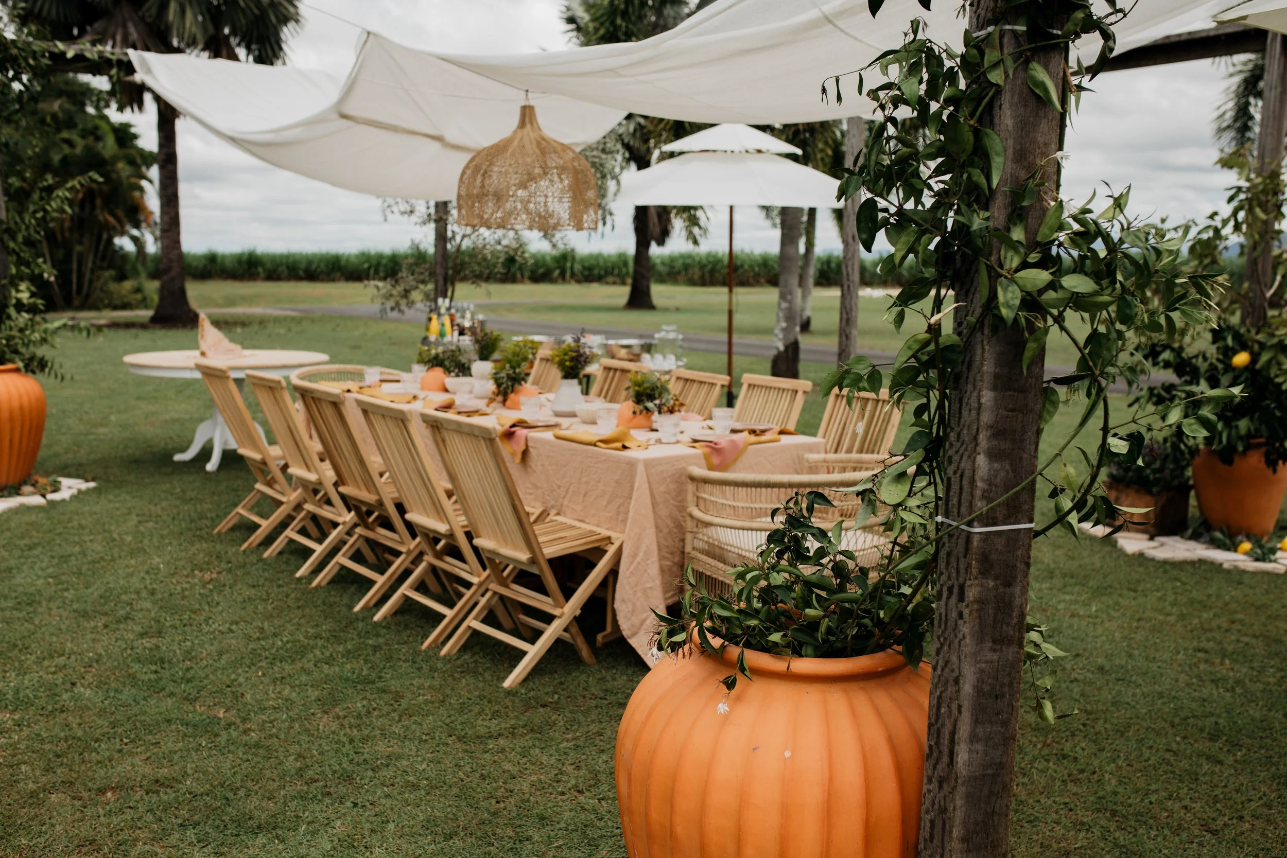 An outdoor dining setup featuring a long wooden table with a beige tablecloth, surrounded by chairs, set under white umbrellas with decorative hanging lights, in a grassy area with trees and large pots filled with greenery.
