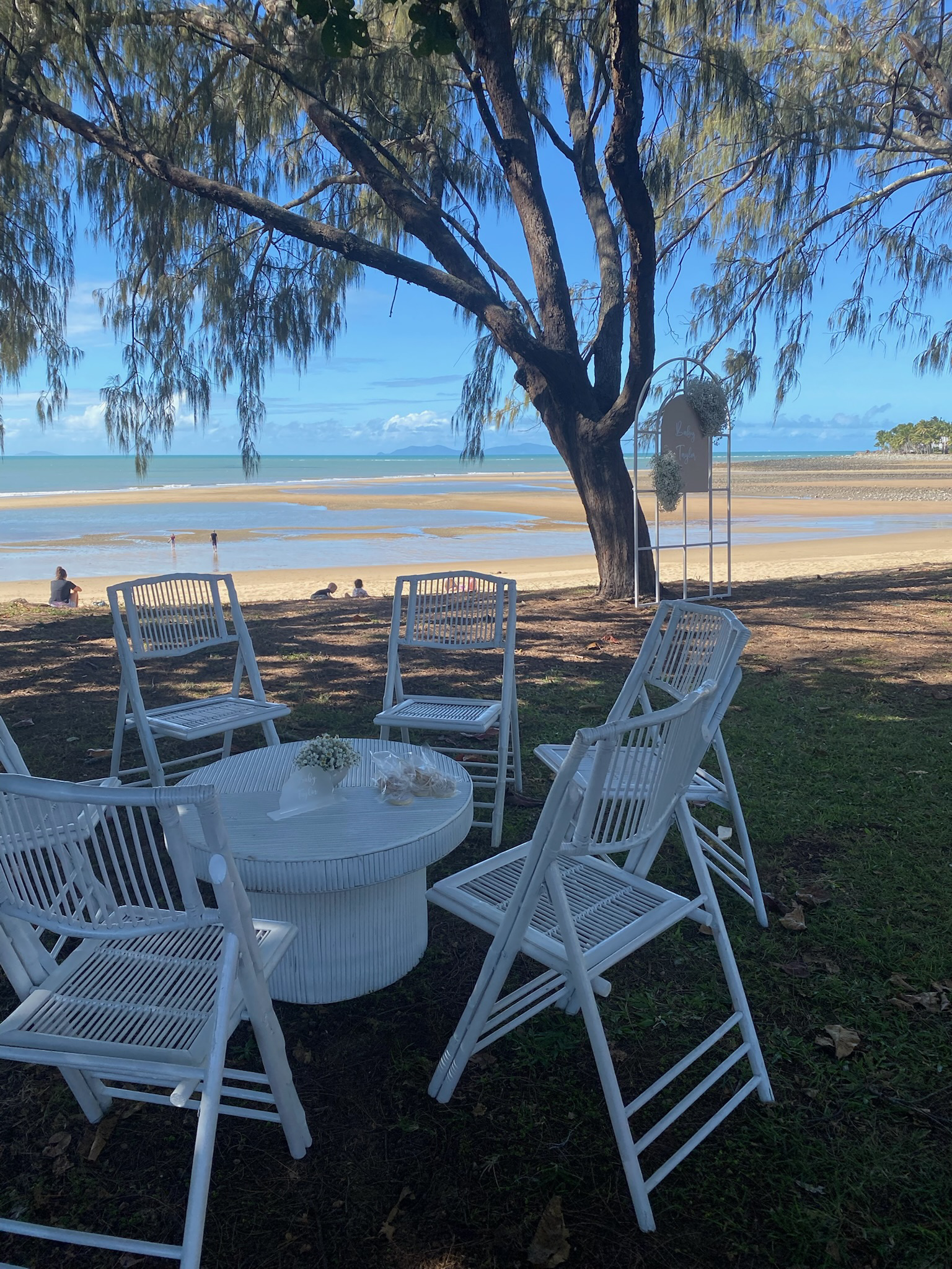 Beachside outdoor setting with white chairs arranged around a white table with flowers and small snacks, under a large tree with a view of the sandy beach and ocean in the background.