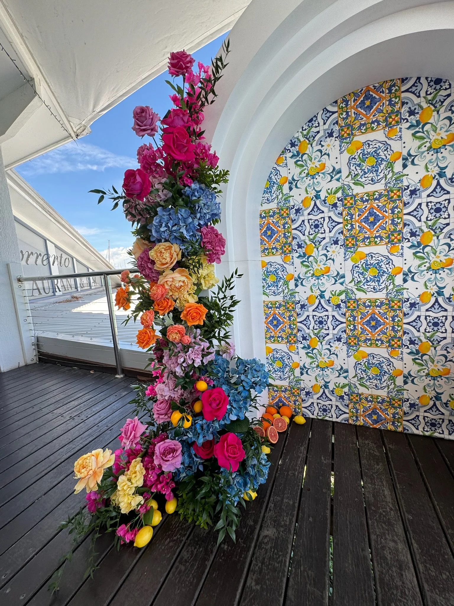 Colorful floral arrangement with pink, purple, blue, yellow, and orange flowers next to a patterned tiled wall under a white arch.