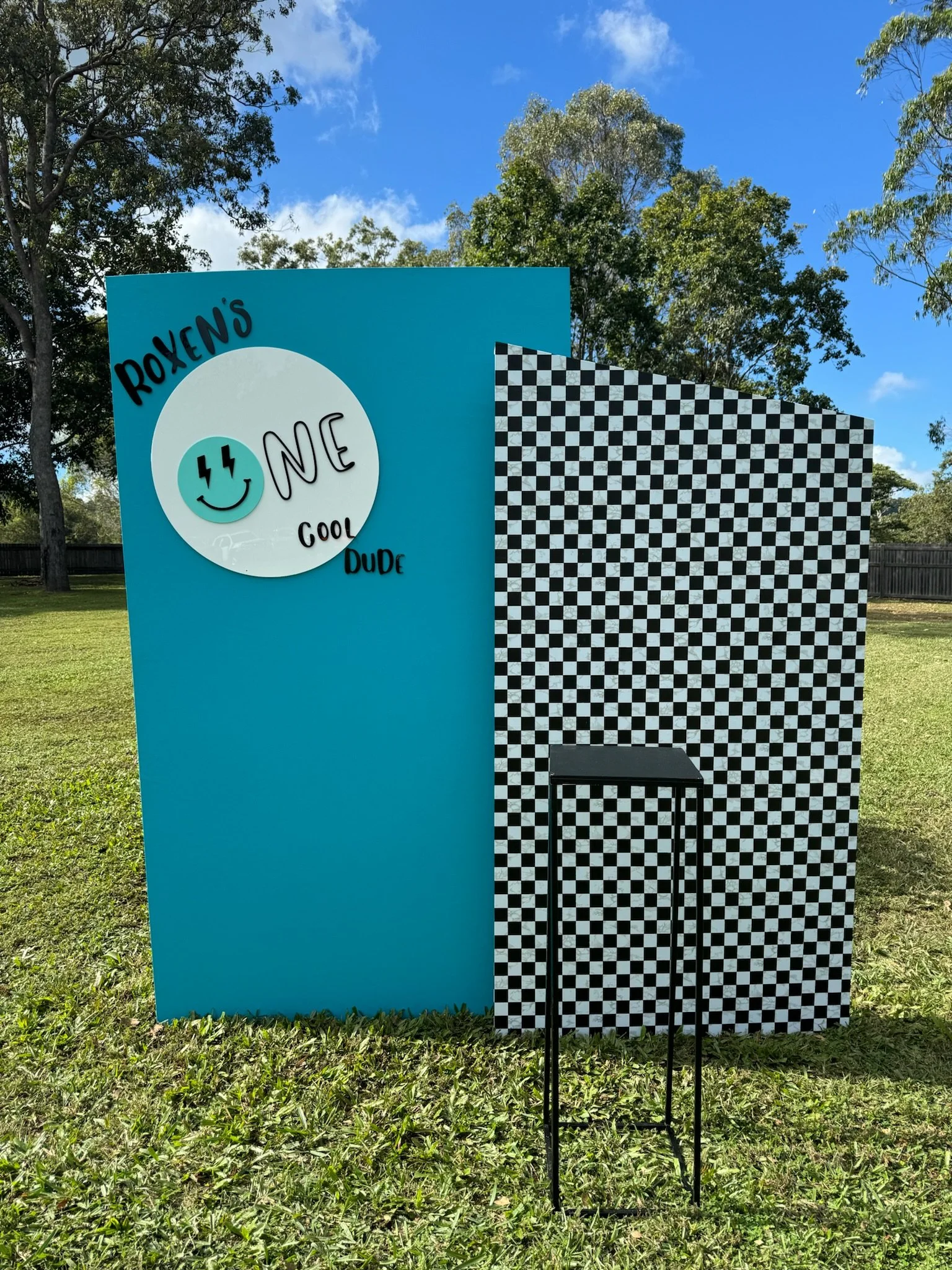 An outdoor photo of a colorful photo booth with a blue wall and black-and-white checkered back wall. The booth has a sign with a smiley face and words like 'Roxen's', 'one', 'cool', 'dude', and 'me' written in playful fonts. There is a small black ta
