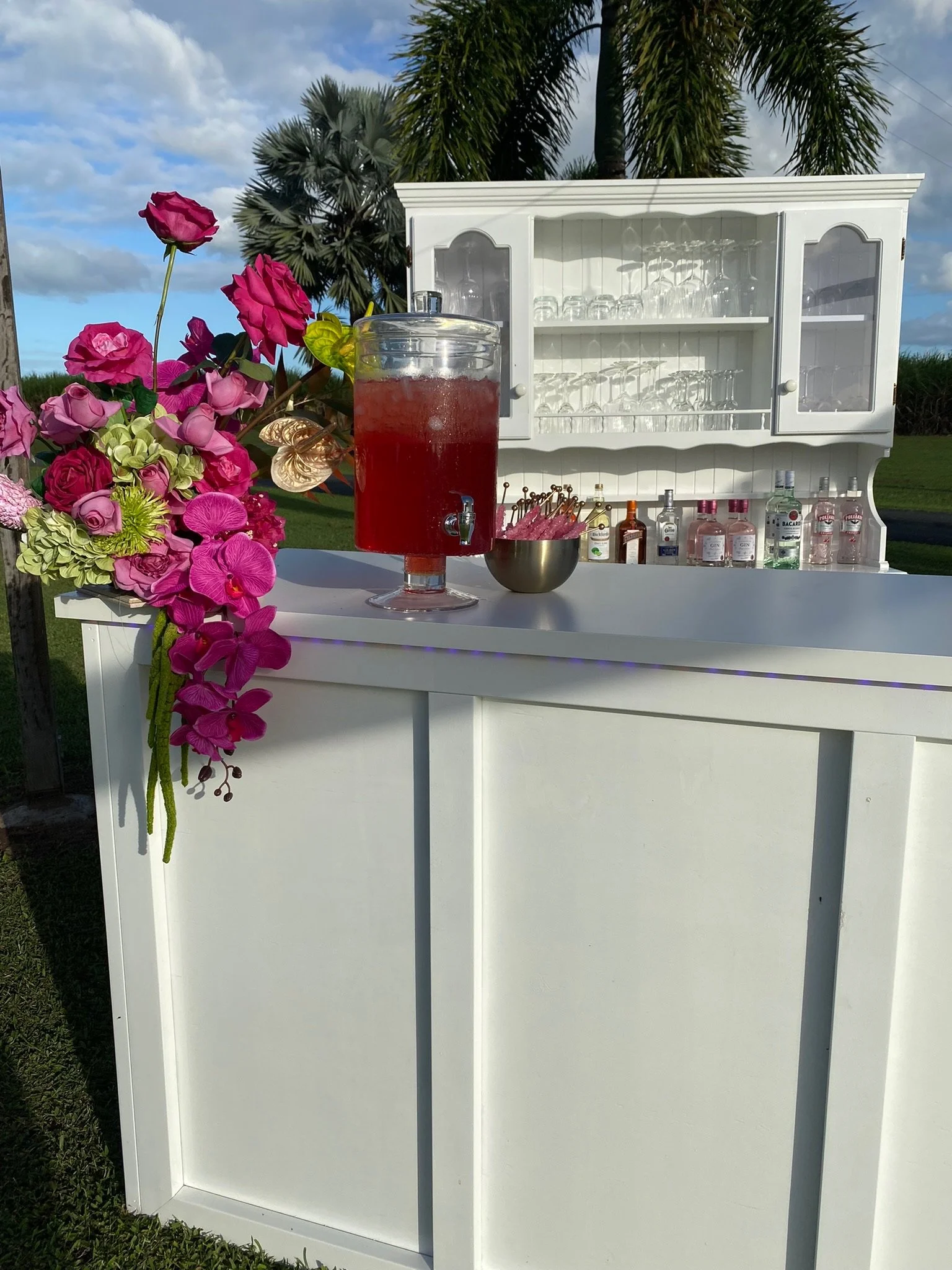 A white outdoor bar setup with pink and purple flowers on the side, a large glass container filled with red drink, and various bottles of alcohol in the background, set against a backdrop of palm trees and a partly cloudy sky.