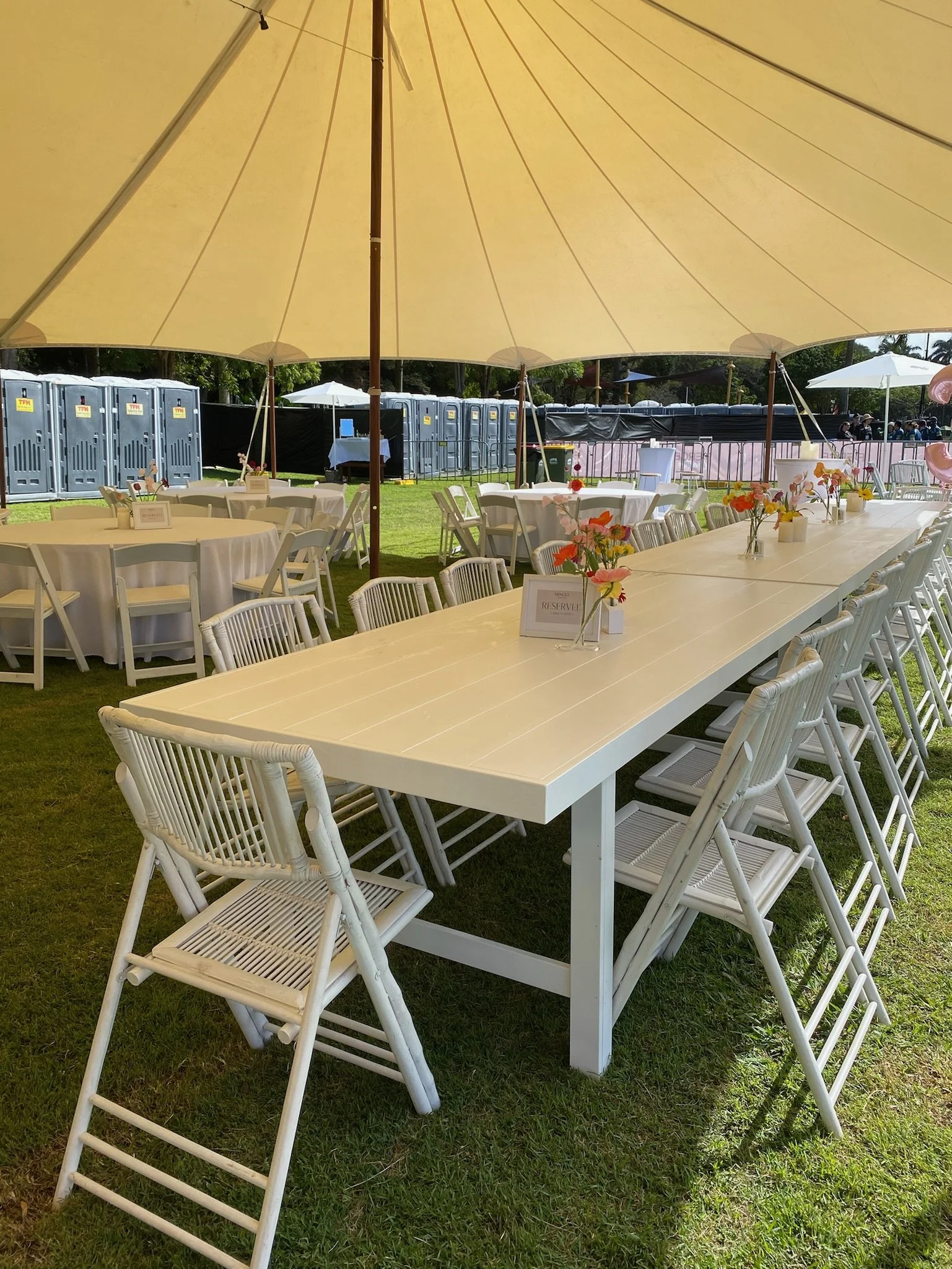 Outdoor event setup with white tables and chairs under a large yellow umbrella, decorated with small flower arrangements, on a grassy area with portable toilets and other seating in the background.