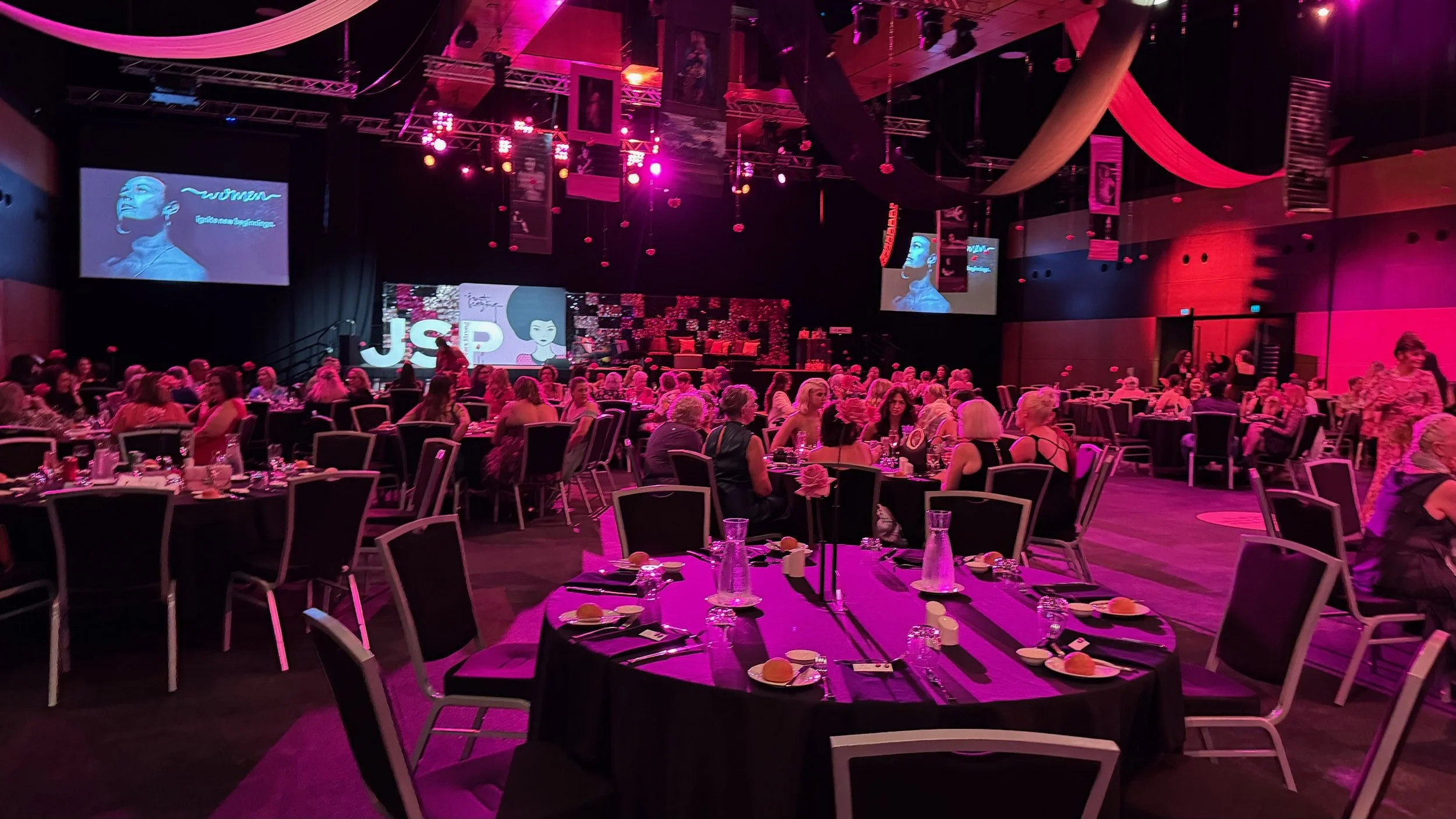 An event hall with round tables set for a formal dinner, decorated with black tablecloths, pink flowers, and glasses, with a stage in the background and people dressed in elegant attire, illuminated with pink lighting.