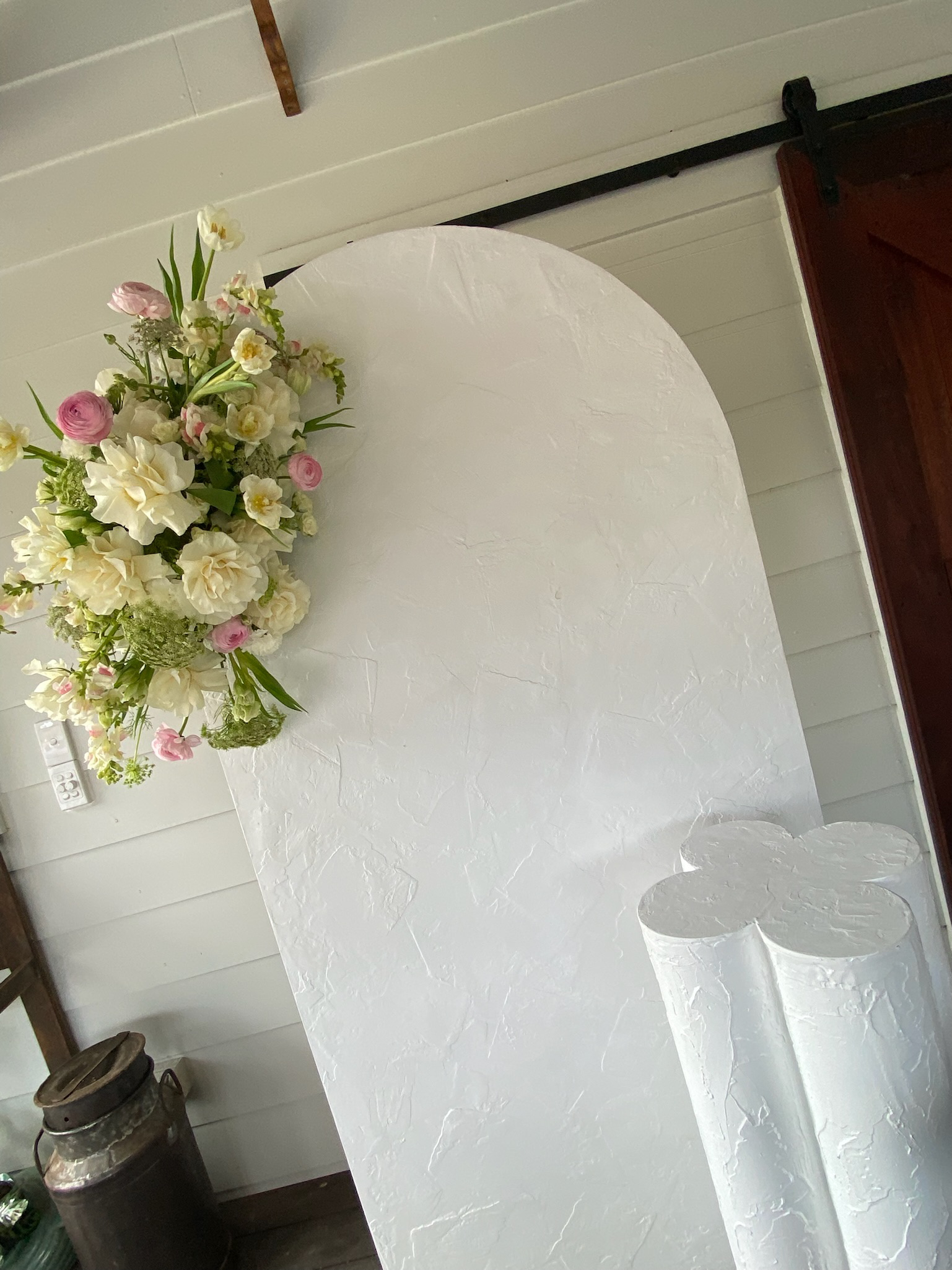 A white textured backdrop with a floral arrangement of white and pink flowers on the left side and three white textured cylinders on the right side. The setting appears to be indoors.