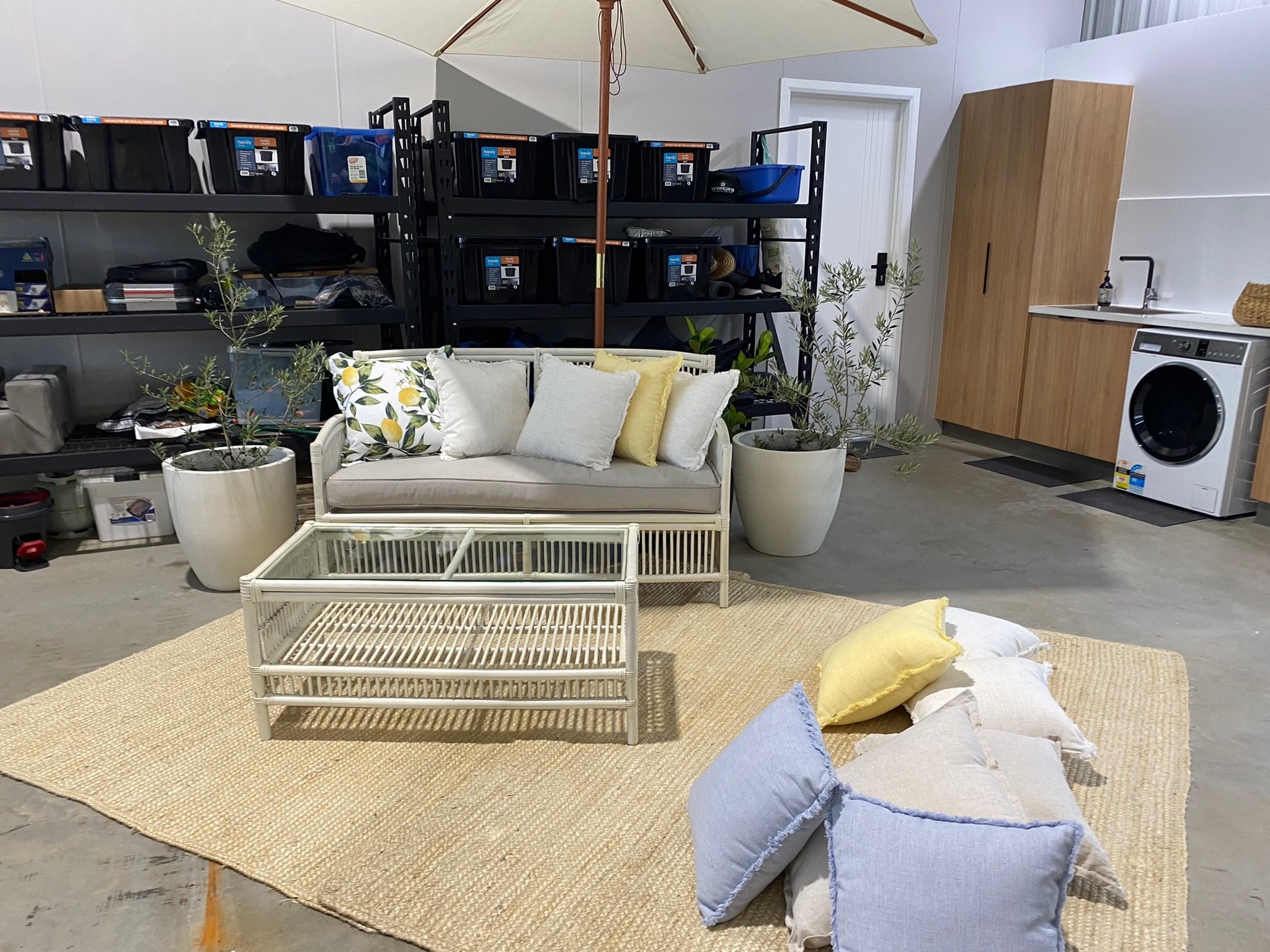 Living space with white sofa, green and white decorative pillows, several scattered pillows on a woven rug, large potted plants, a glass coffee table, black shelving with storage boxes, a washing machine, a wooden cabinet, and a laundry sink in the b