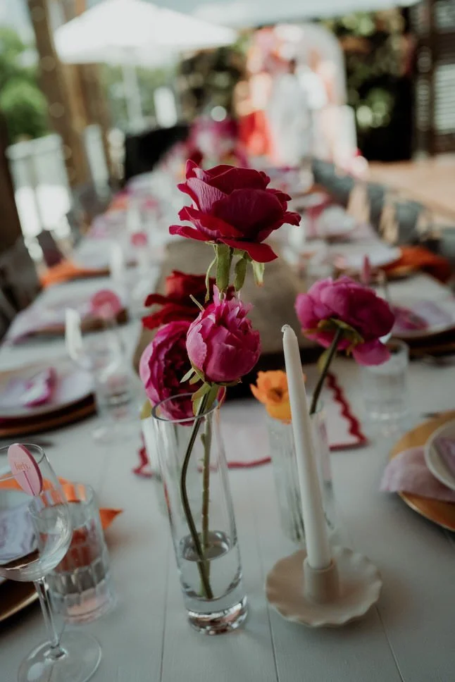 Close-up of pink and red flowers in a tall glass vase on a dining table set for an event.