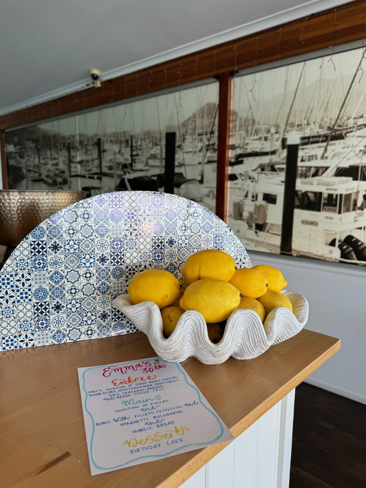 A basket of lemons on a wooden counter with a decorative blue and white patterned fan and a handwritten menu, in front of a mural of boats docked at a marina.