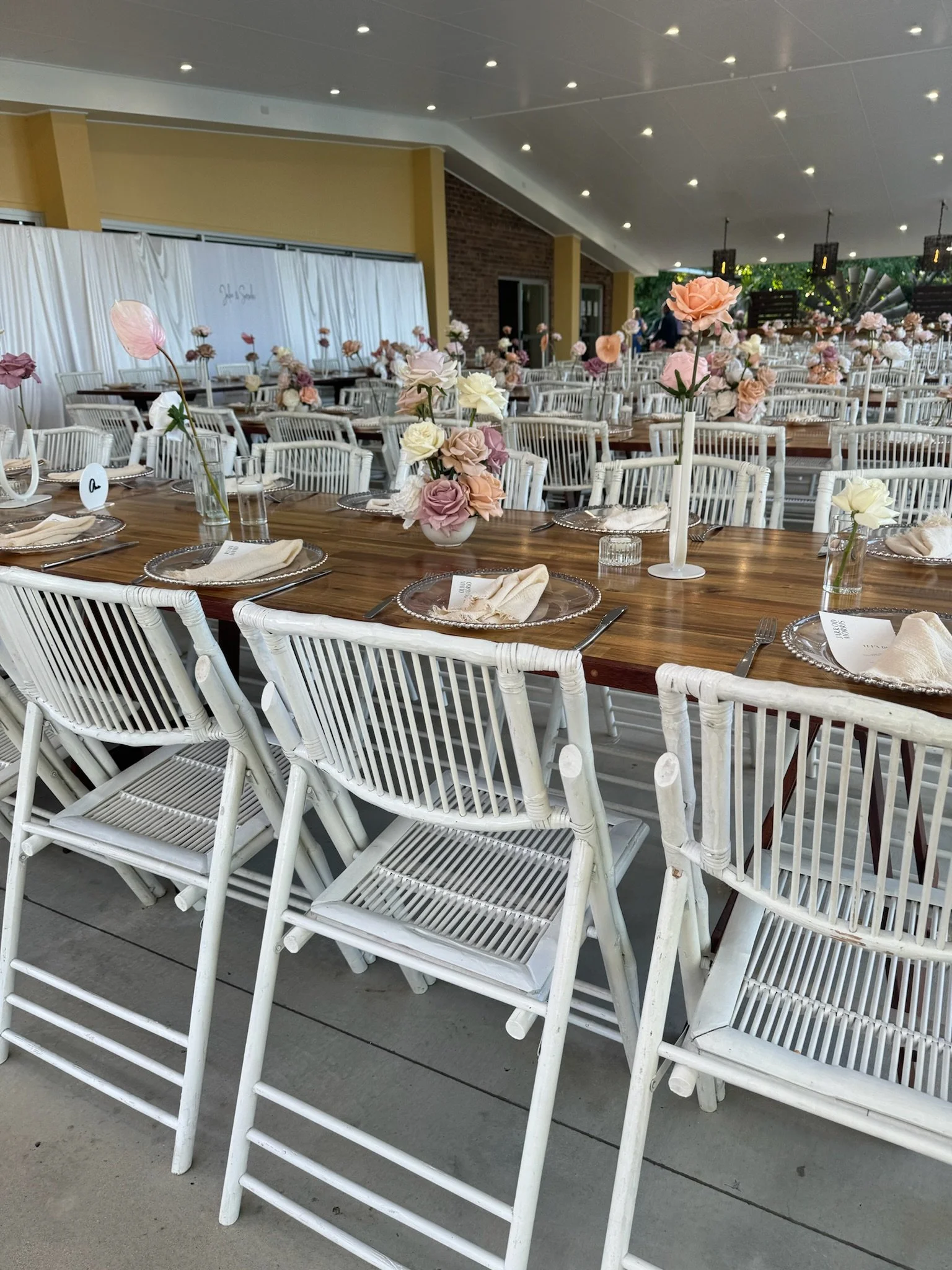 Wedding reception setup with long wooden tables, white wicker chairs, and floral centerpieces with pink, white, and blush roses, inside a modern hall with a yellow wall, brick accents, and a white draped backdrop.
