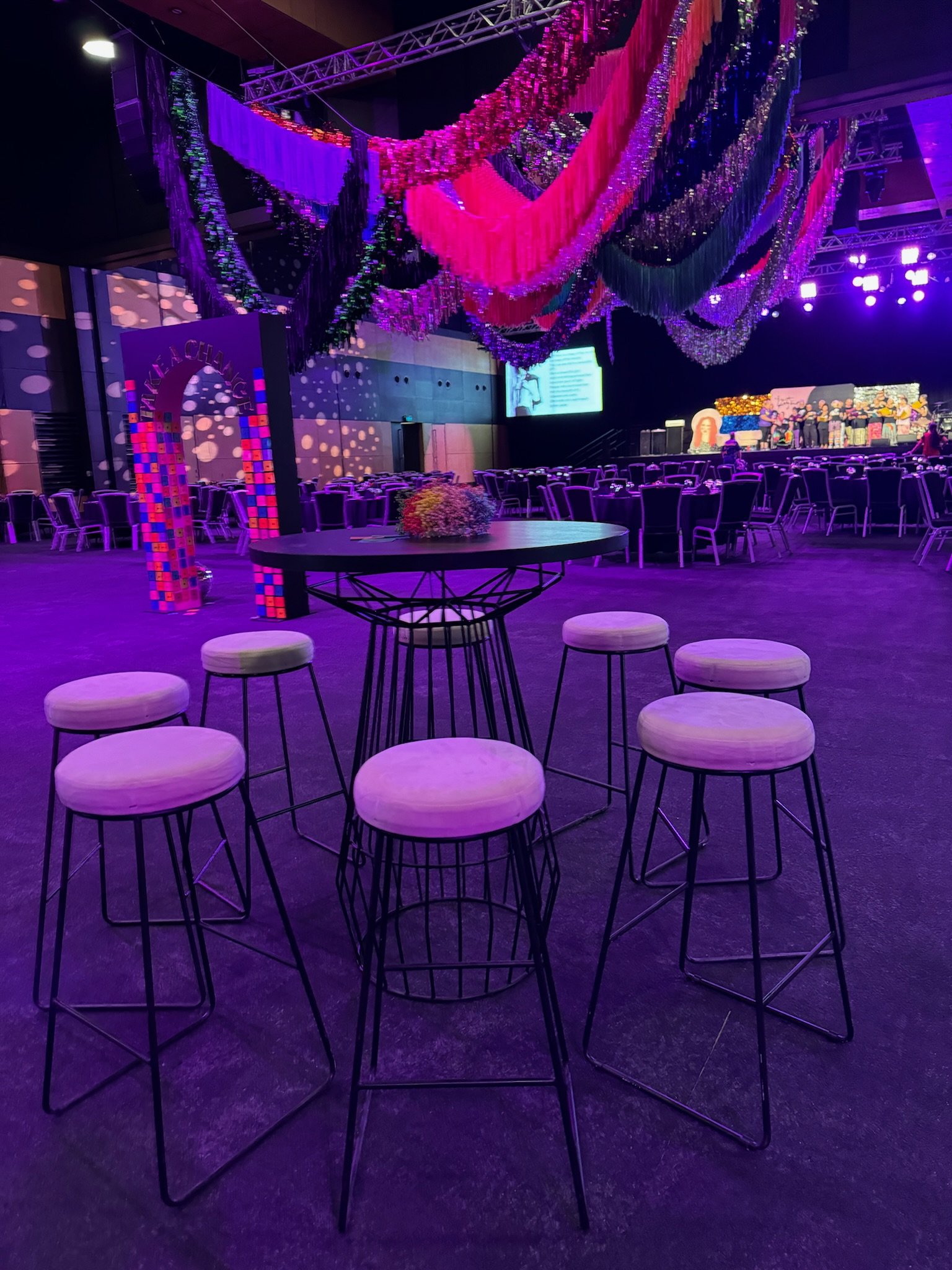 Interior of a decorated event or conference hall with purple and pink lighting, hanging streamers and chains, and round tables and chairs.