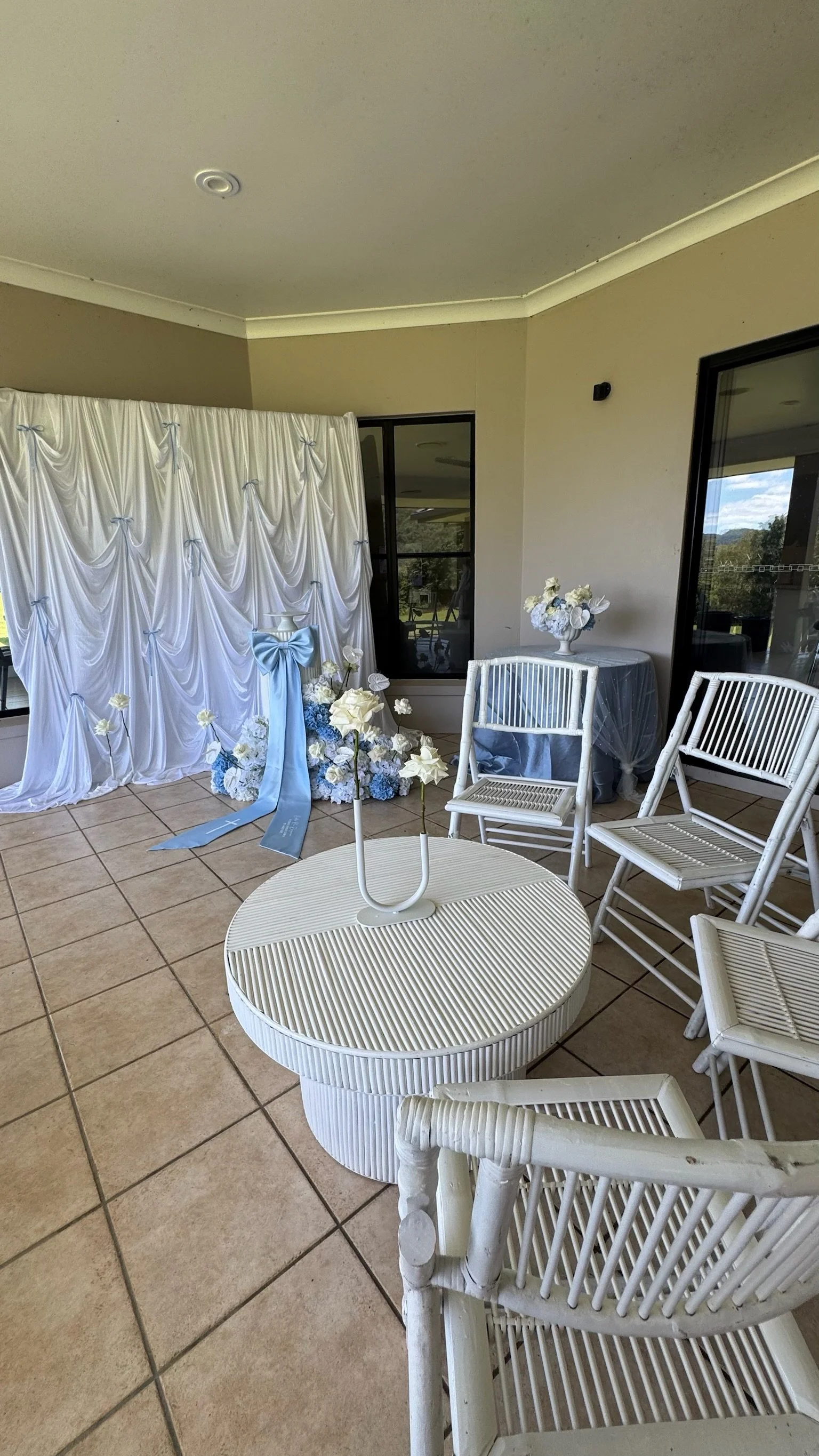 Decorative white backdrop with blue ribbons, floral arrangements including white roses and blue hydrangeas, set up on a patio with white chairs and round tables.