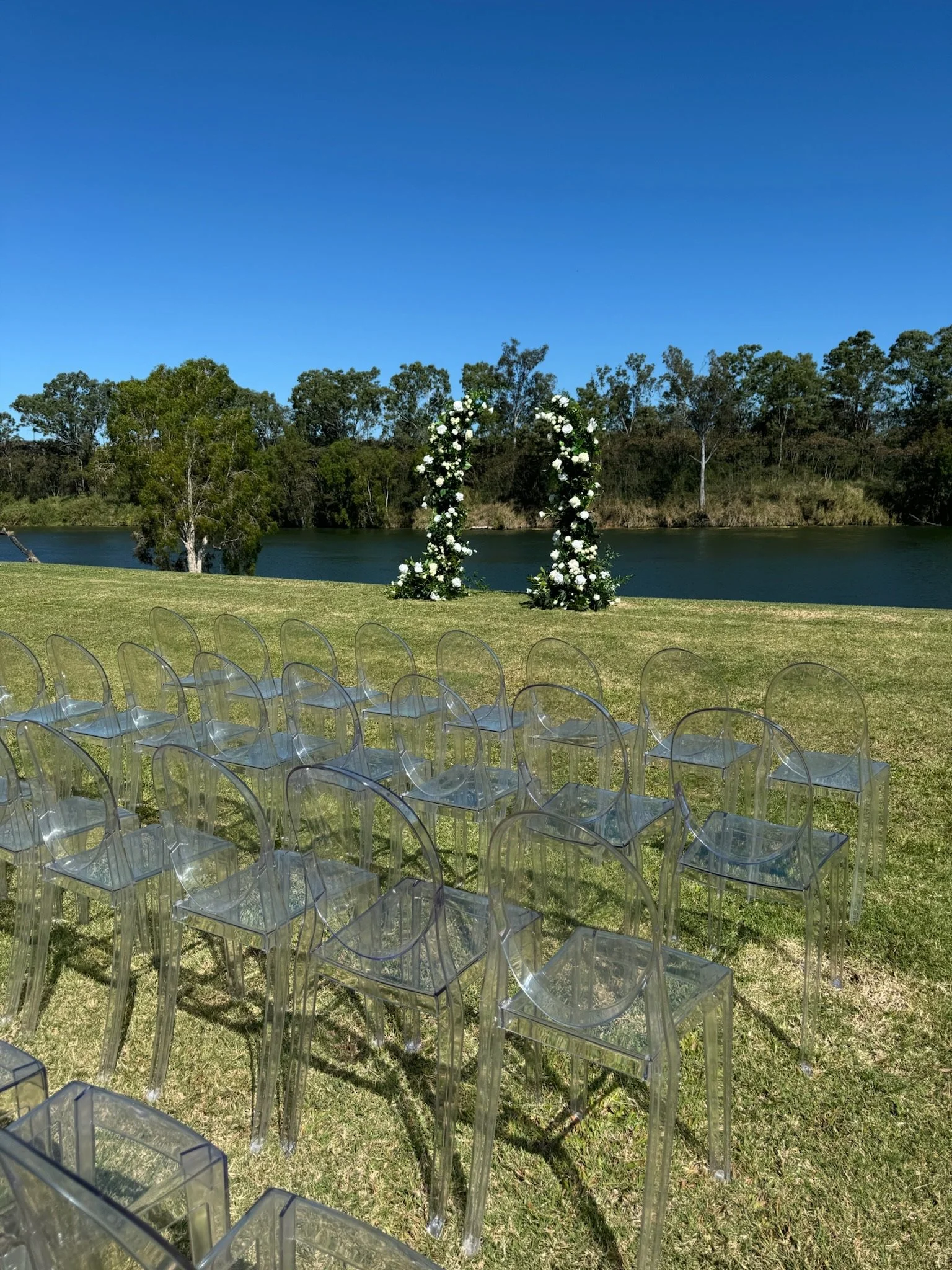 Outdoor wedding setup with clear chairs arranged in rows on grass facing a floral arch by a lake under a clear blue sky.