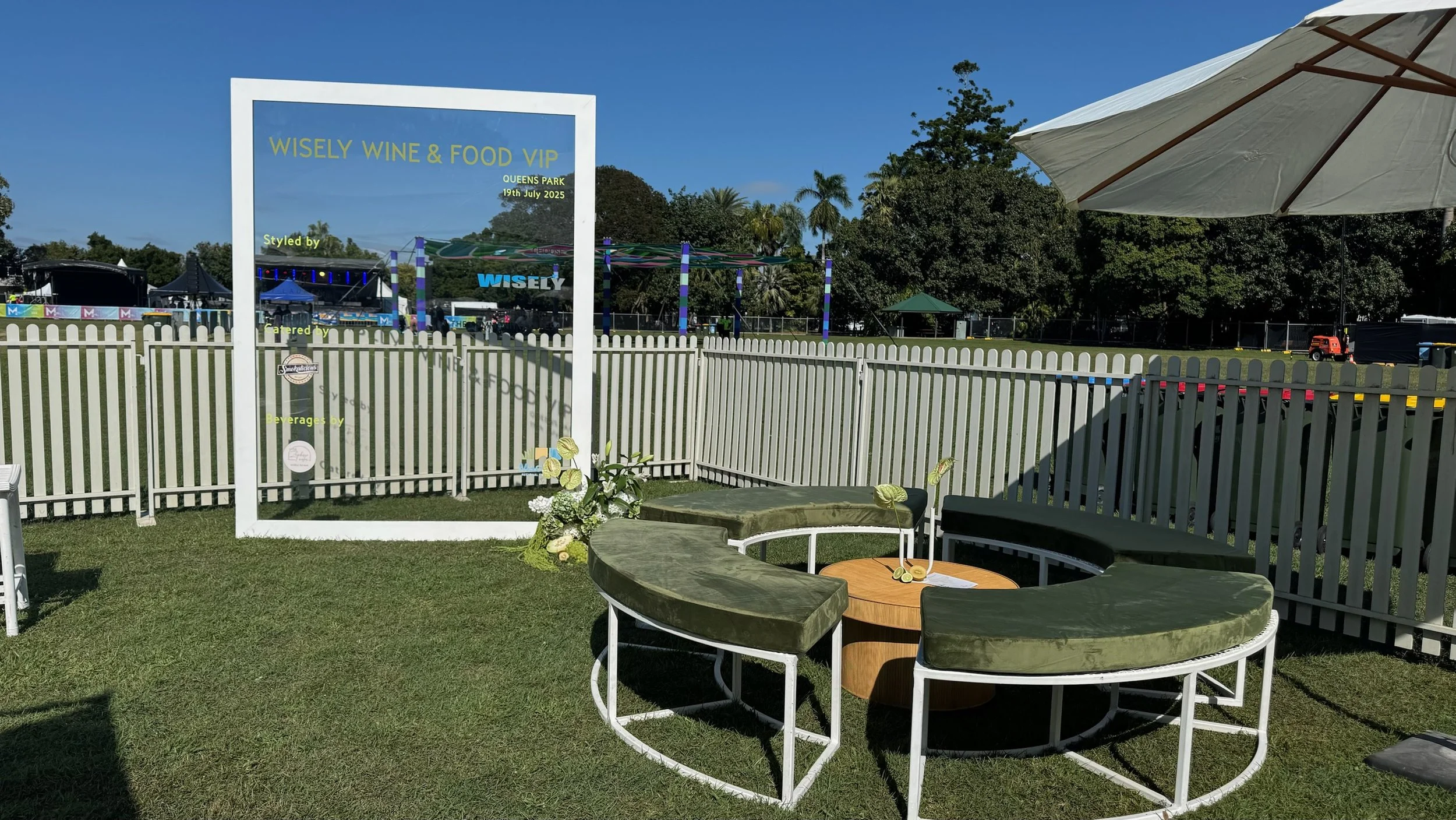Outdoor event setup with white picket fence, circular green velvet seating, a small round wooden table, floral decoration, and a large white frame with some text, at Wise Wine & Food VIP event at Queens Park on July 19, 2025, with amusement park ride