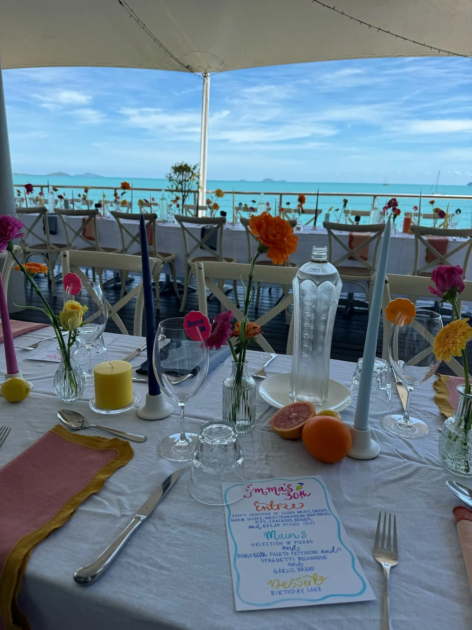 A beautifully set seaside outdoor dining table decorated with colorful flowers, candles, and a menu, overlooking the ocean with islands in the distance under a blue sky.