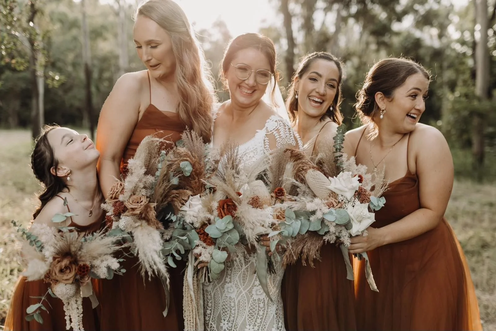 A group of women and a girl smiling and holding bouquets outdoors during daytime, with trees in the background.