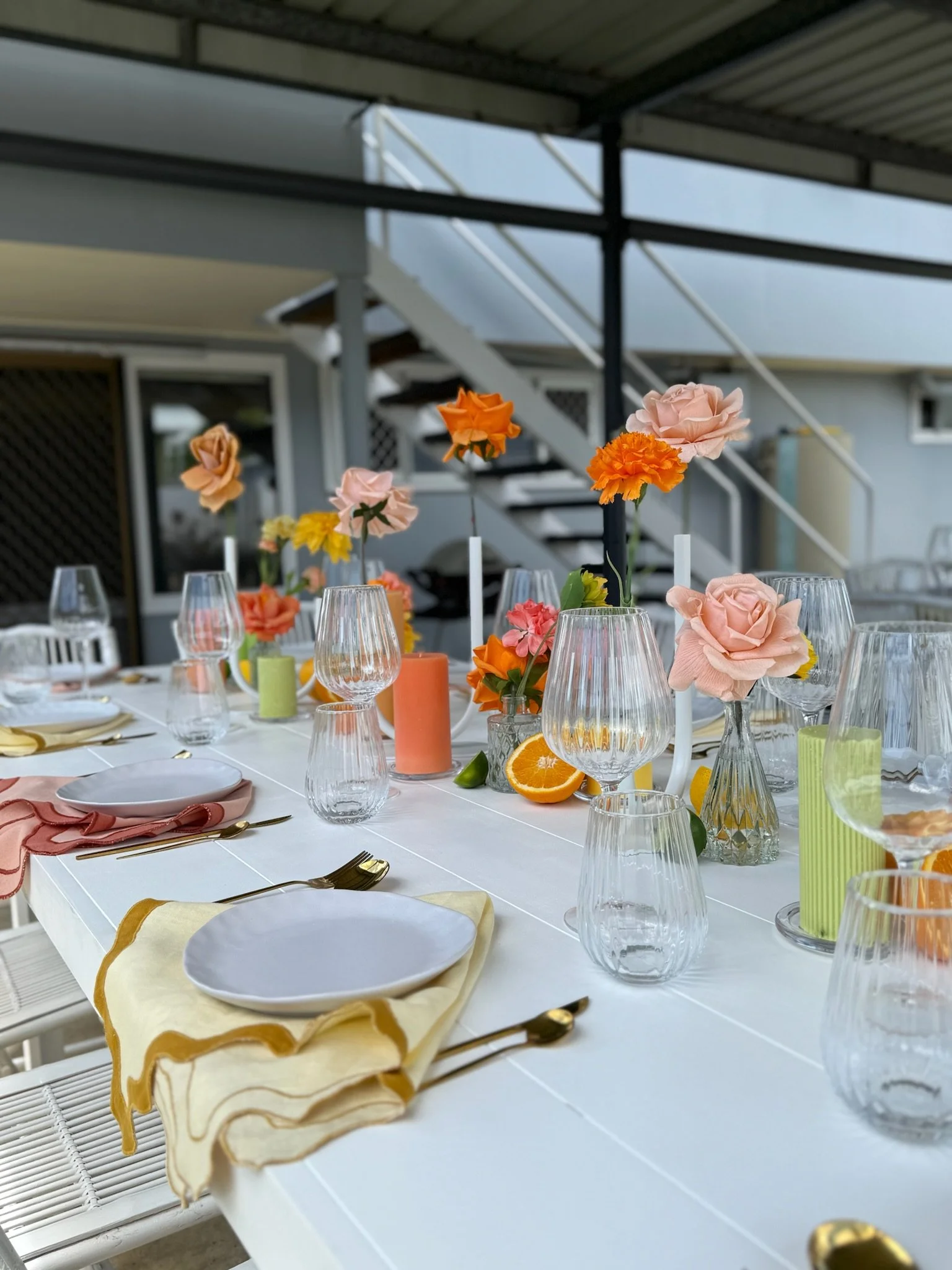 Set dining table with pink, orange, yellow, and peach flowers in vases, lit candles, glasses, plates, cutlery, and napkins in pastel colors, outdoors on a patio with a staircase in the background.