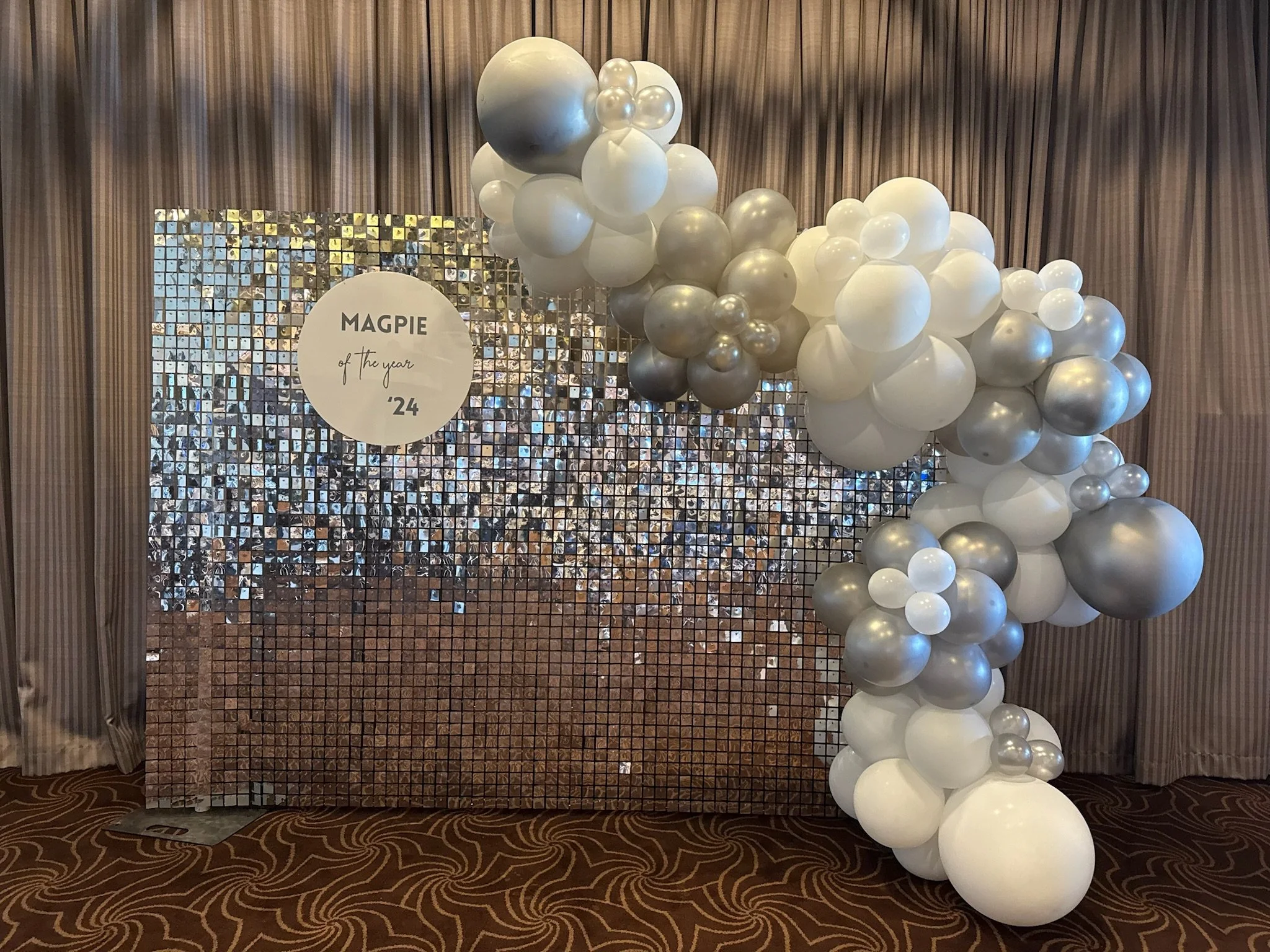 Decorative backdrop with a cluster of white, silver, and pearl balloons on the right side, and a shimmering, mosaic-style surface with a circular sign that reads "Magpie of the year ’24" on the left side, set against brown curtains and patterned carp