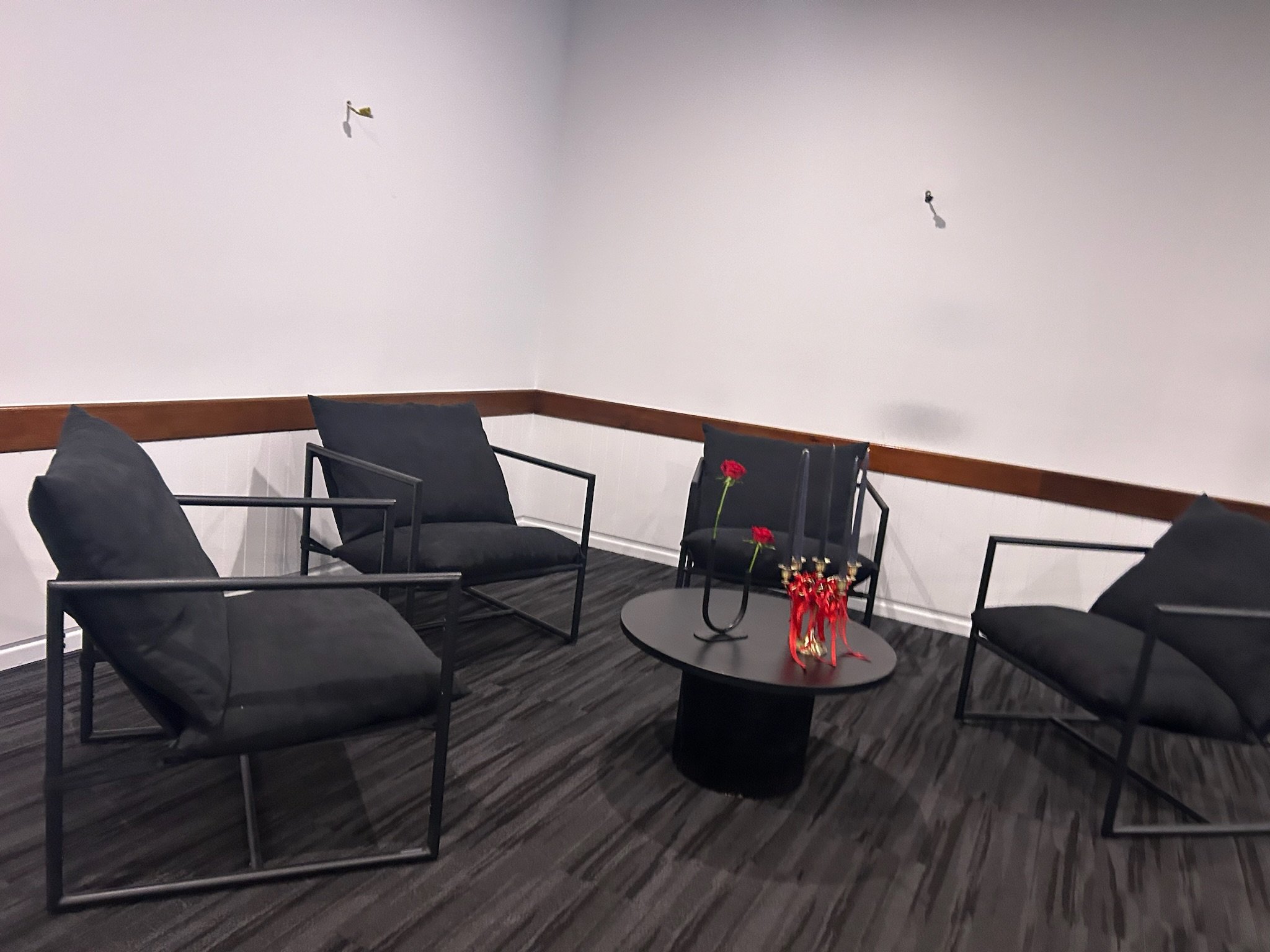 Minimalist seating area with four black armchairs arranged around a black round coffee table, which holds a red vase with red flowers; plain white walls with wood trim, wood flooring, and two wall-mounted lights.