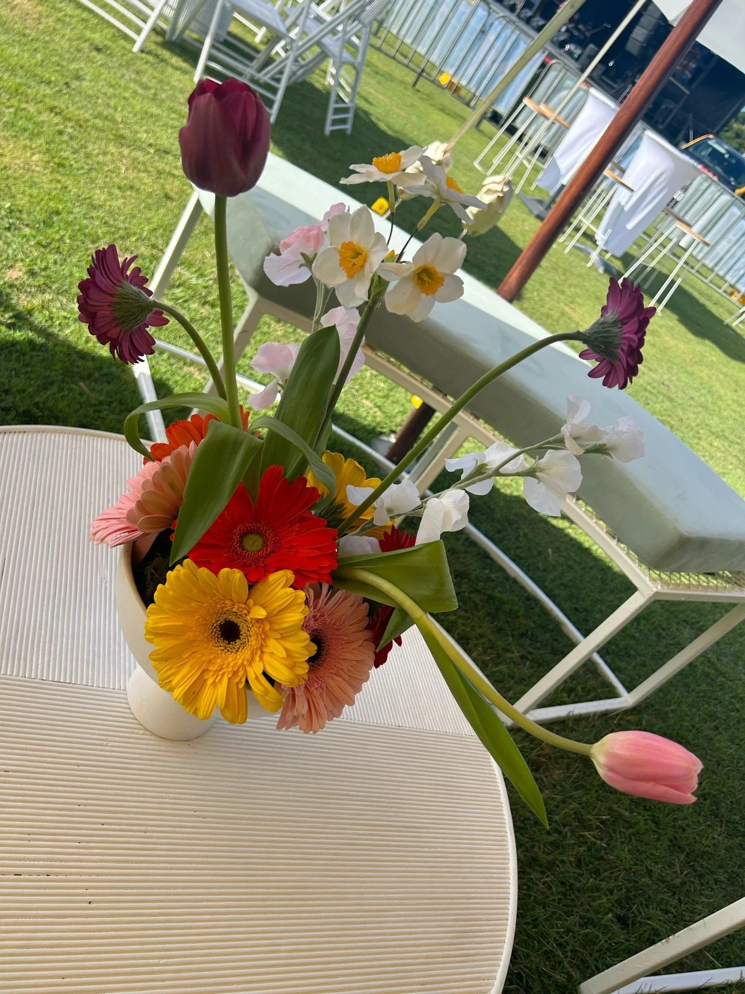 A colorful flower arrangement in a white vase on a white outdoor table, including tulips, daisies, gerbera daisies, and primroses, with a grassy area and tables with white cloths in the background.