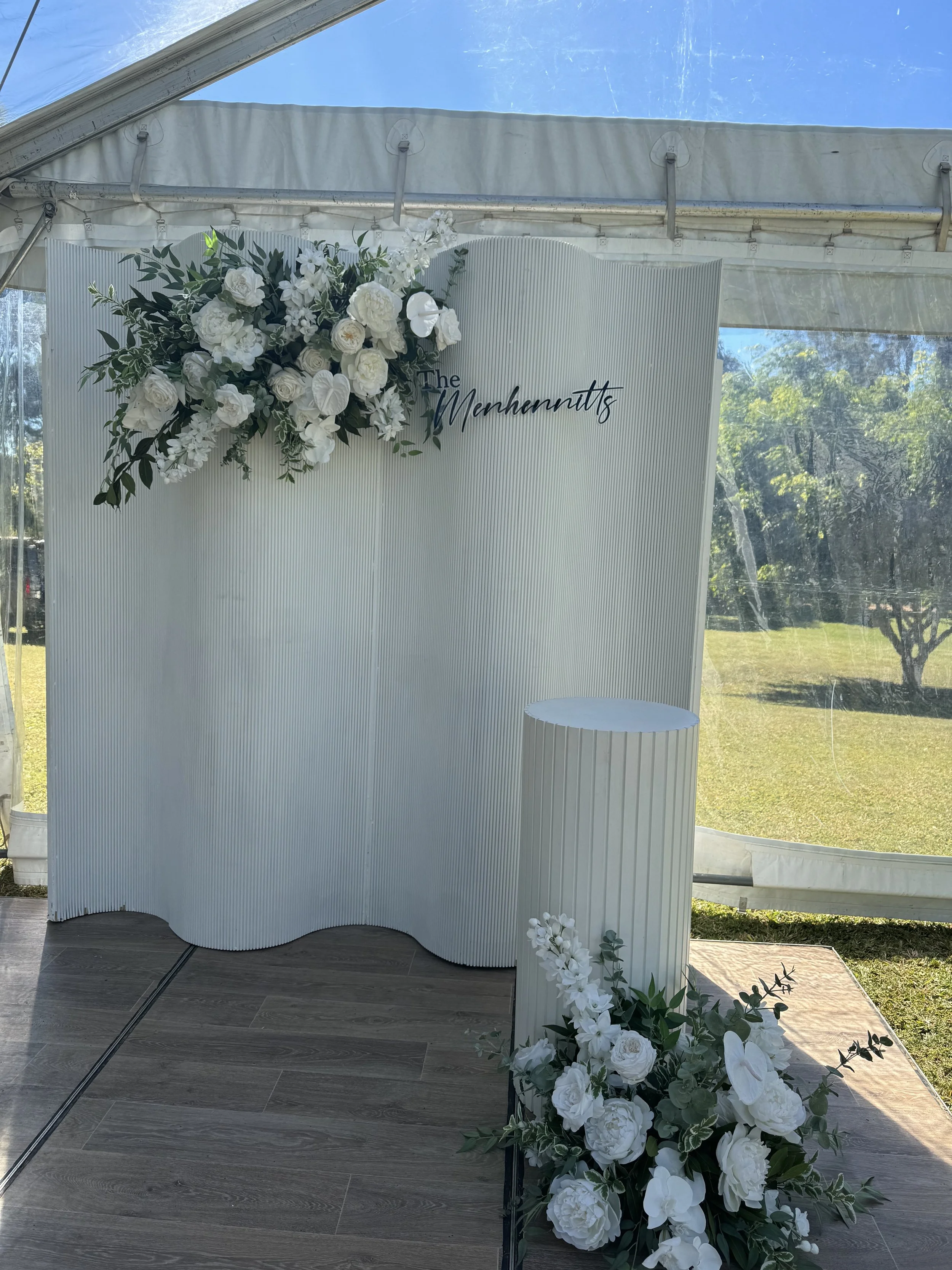 Elegant wedding or event backdrop with white floral arrangements, a white textured curved partition, and a sign that reads 'The Merchants' in black script, set up outdoors under a tent with greenery in the background.